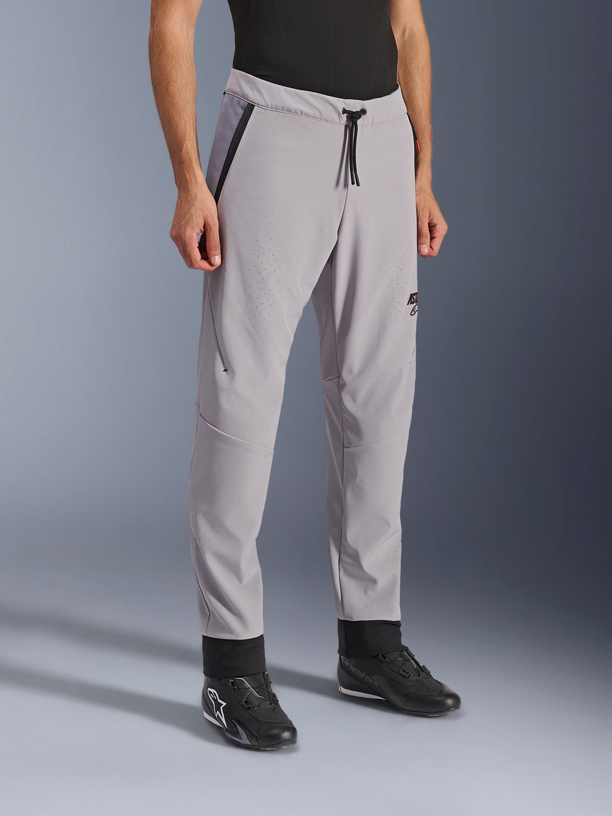 Pantalon Alpinestars Aeroshell Airflow, gris moyen, pantalon de moto léger avec zones de ventilation perforées au laser sur les cuisses et les tibias, doté d'une taille élastique avec cordon de serrage et d'une coupe sport ajustée, porté par un mannequin avec des chaussures de moto noires.