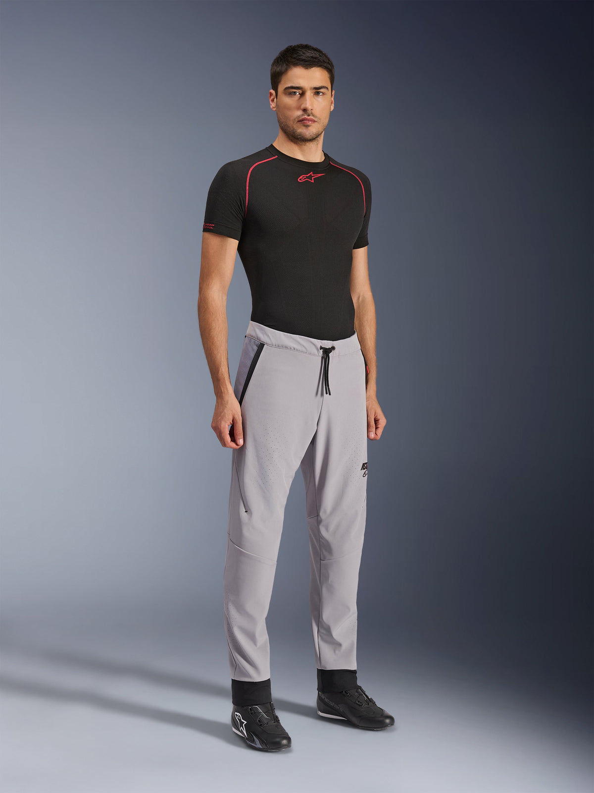 Pantalon Aeroshell Airflow Alpinestars, pantalon de moto, gris moyen, porté par un mannequin avec un t-shirt technique noir et des chaussures de moto noires, doté de zones de ventilation perforées au laser, d'une taille élastique avec cordon de serrage et d'une coupe sportive profilée.