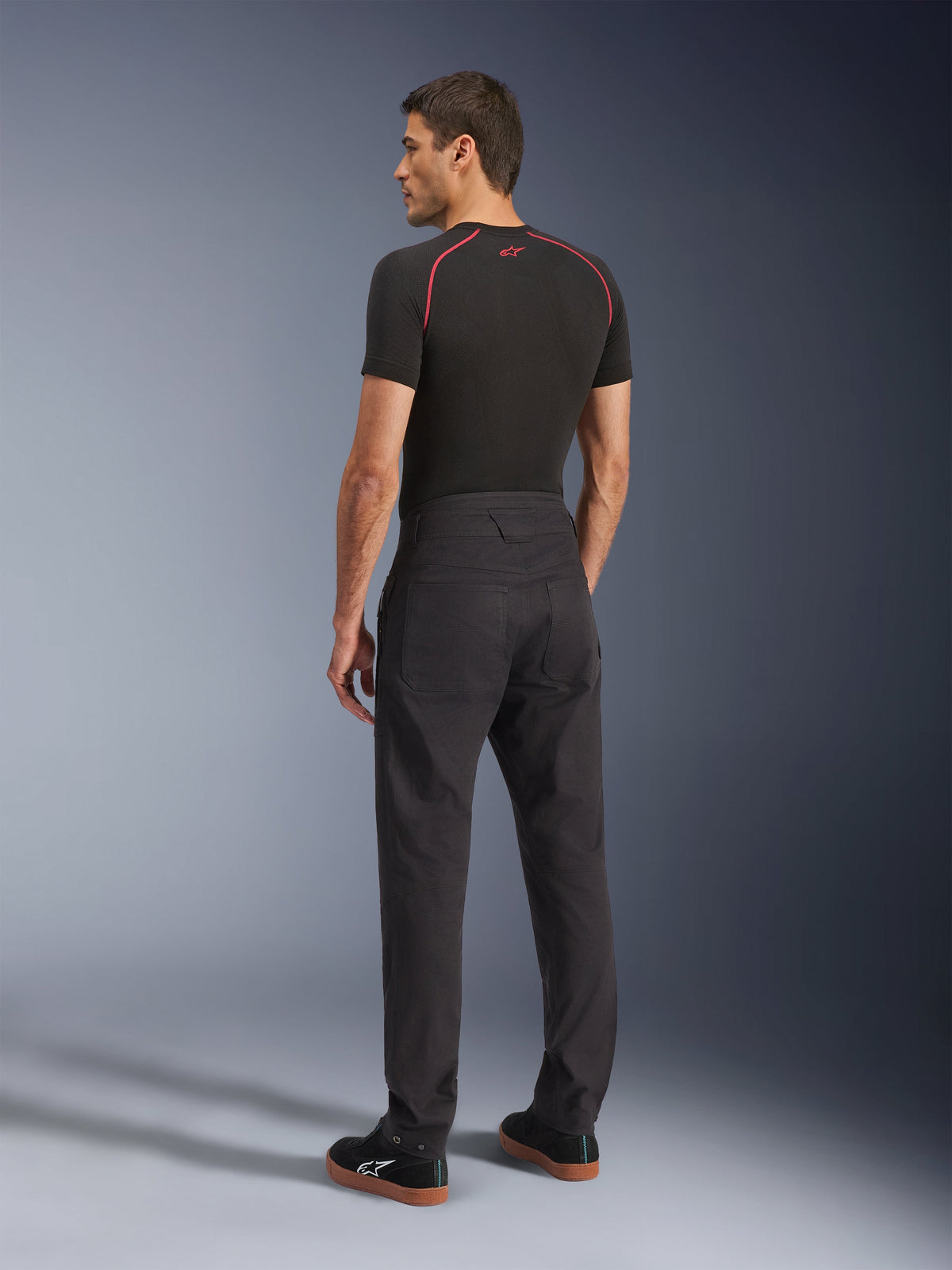 Pantalon Flex-AST Canvas Alpinestars, pantalon de moto urbain noir, vue arrière sur modèle, associé à une couche de base noire à manches courtes et des baskets noires, doté d'un design inspiré du style workwear avec de grandes poches arrière et une coupe droite moderne.