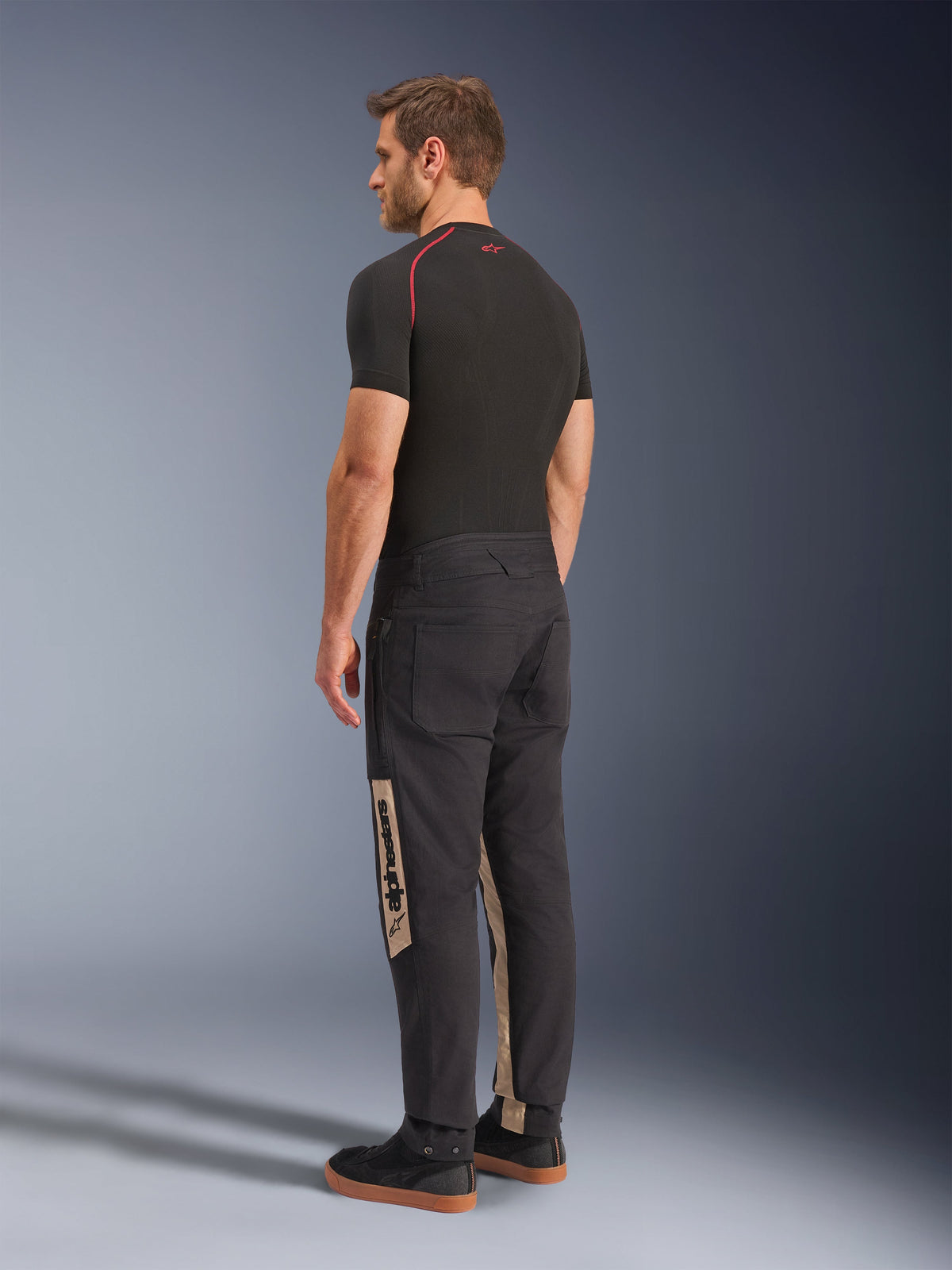 Pantalon Alpinestars Flex-AST Canvas, Black Aluminum, pantalon de moto urbain, avec empiècements latéraux beiges et marquage Alpinestars, porté avec une couche de base noire et des baskets, vue arrière montrant un style inspiré du workwear et une coupe moto ergonomique.