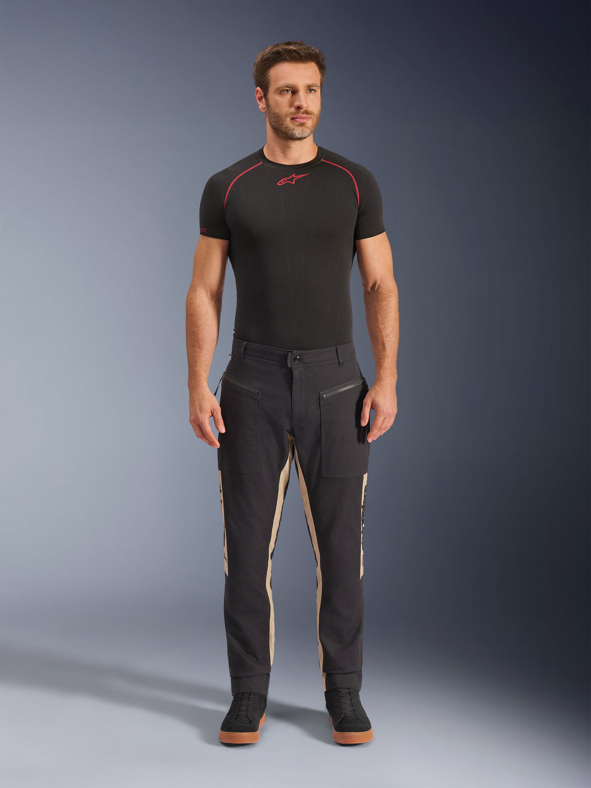 Modèle masculin portant le pantalon Alpinestars Flex-AST Canvas en Noir Aluminium, pantalon de moto urbain, noir avec panneaux beiges, doté de poches utilitaires et de fermetures éclair d'inspiration workwear, associé à une couche de base noire et des baskets noires