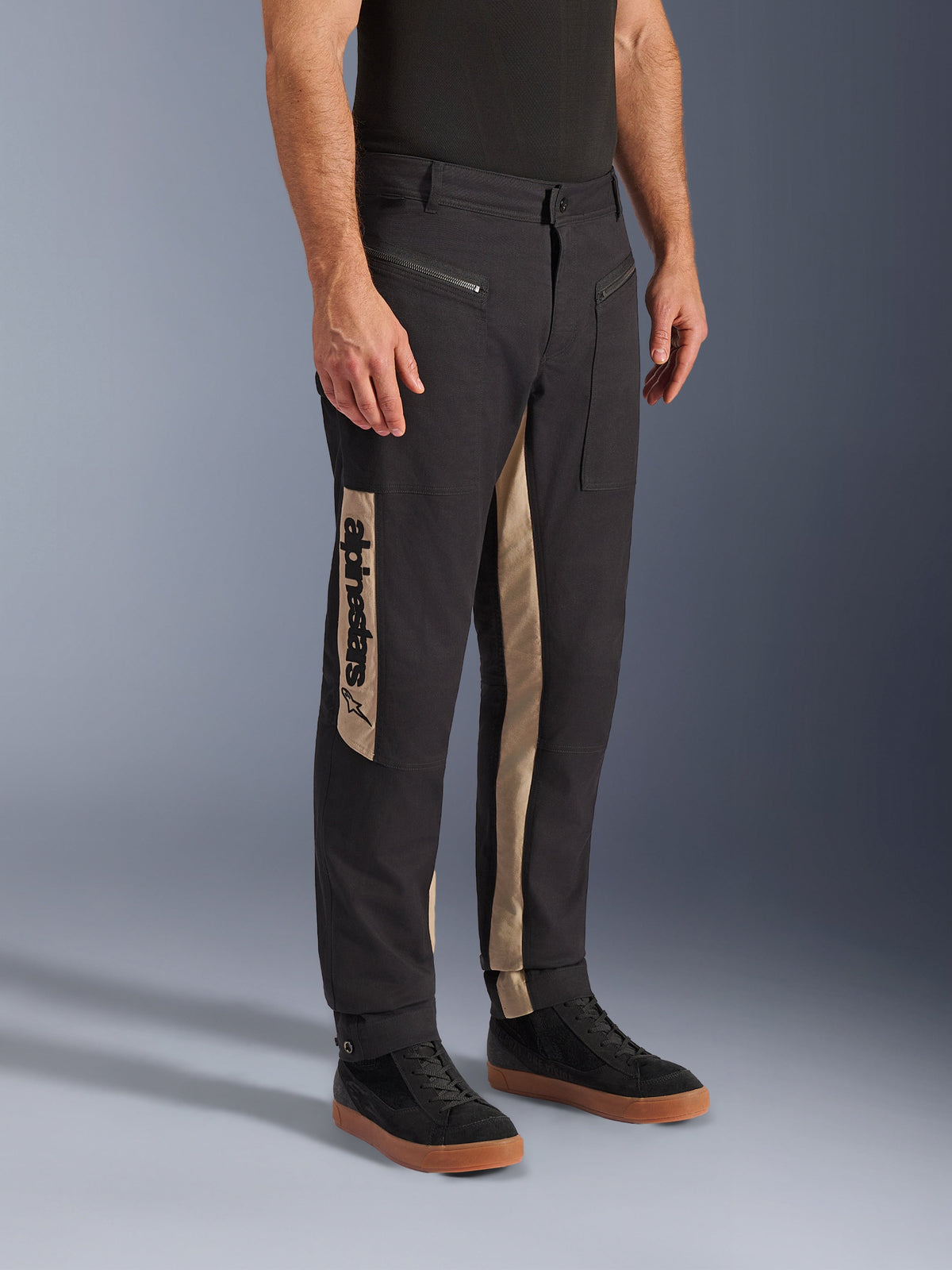 Pantalon Alpinestars Flex-AST Canvas, pantalon de moto urbain, Black Aluminum, doté de panneaux latéraux beiges avec un logo Alpinestars vertical, design inspiré du workwear avec poches zippées et coupe ergonomique, porté avec des baskets noires décontractées pour un look urbain moderne.