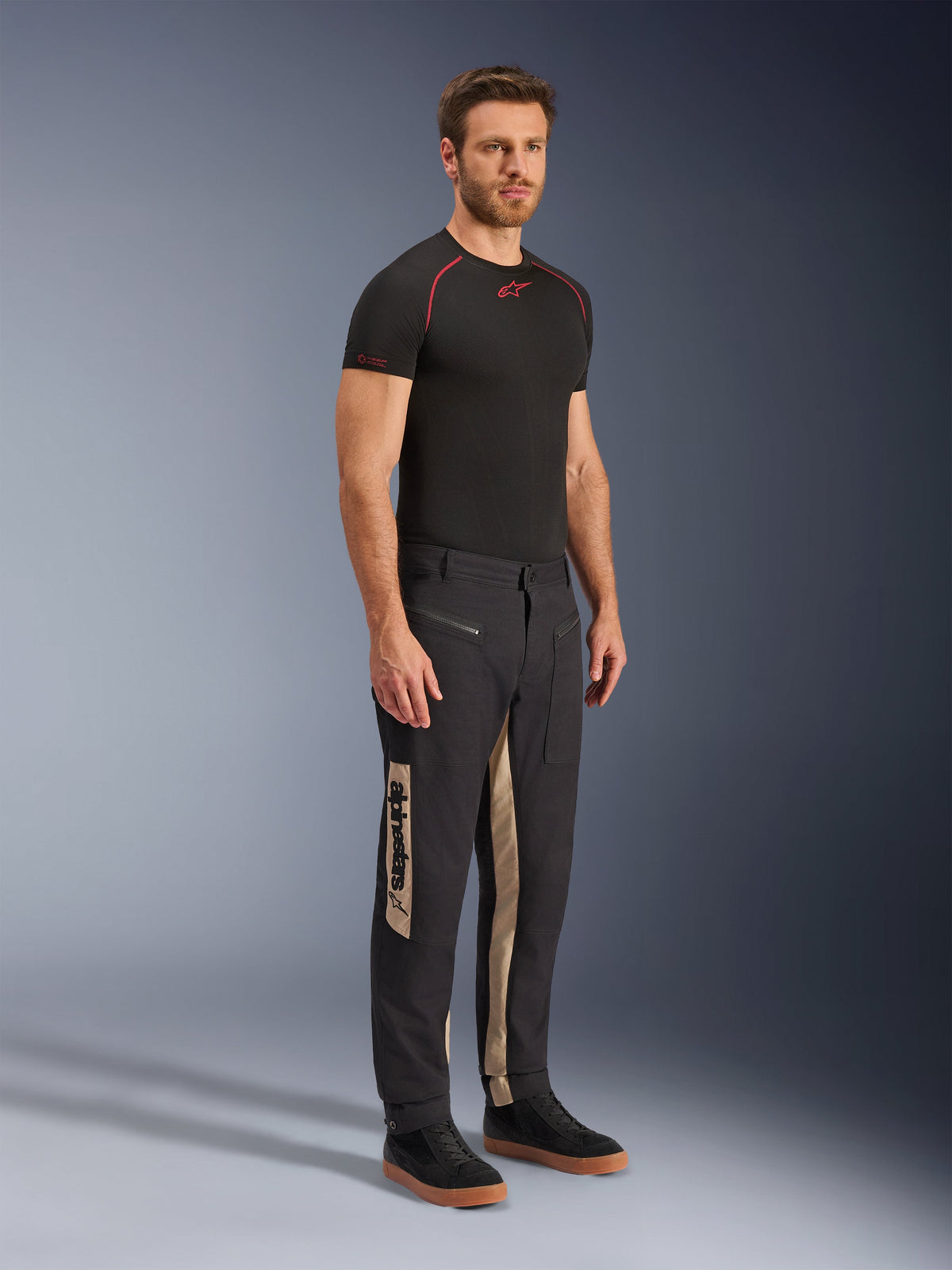 Pantalon Alpinestars Flex-AST Canvas, pantalon de moto urbain, Black Aluminum, porté avec une couche de base technique noire et des baskets noires, doté de panneaux latéraux beiges avec marquage Alpinestars vertical, design utilitaire inspiré du workwear avec genoux préformés et fermetures éclair métalliques