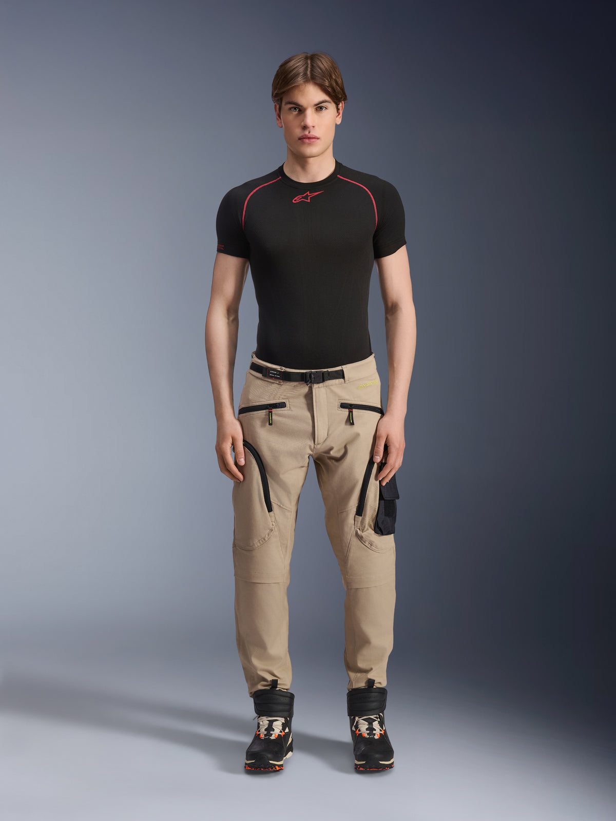 Mannequin homme portant le pantalon Alpinestars Acteon en coloris Sand, associé à une couche de base noire à manches courtes et des bottes techniques noires, doté de poches cargo avec fermetures éclair noires et d'une coupe fuselée, équipement de moto urbain contemporain