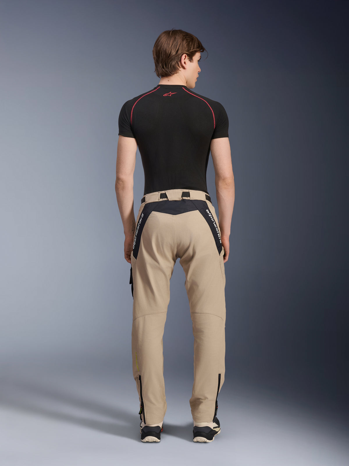 Pantalon Alpinestars Acteon en Sand, pantalon de moto urbain moderne vu de dos, avec empiècements noirs et marquage Alpinestars blanc, coupe fuselée avec fermetures éclair aux chevilles, porté avec une couche de base noire à manches courtes et des baskets.