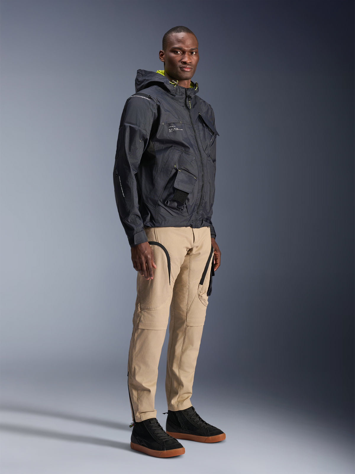 Pantalon Alpinestars Acteon, pantalon de moto urbain, couleur sable, porté par un mannequin avec une veste technique sombre et des baskets noires, doté d'une coupe fuselée avec des détails de fermetures éclair noires et des poches cargo, design contemporain inspiré des sports mécaniques