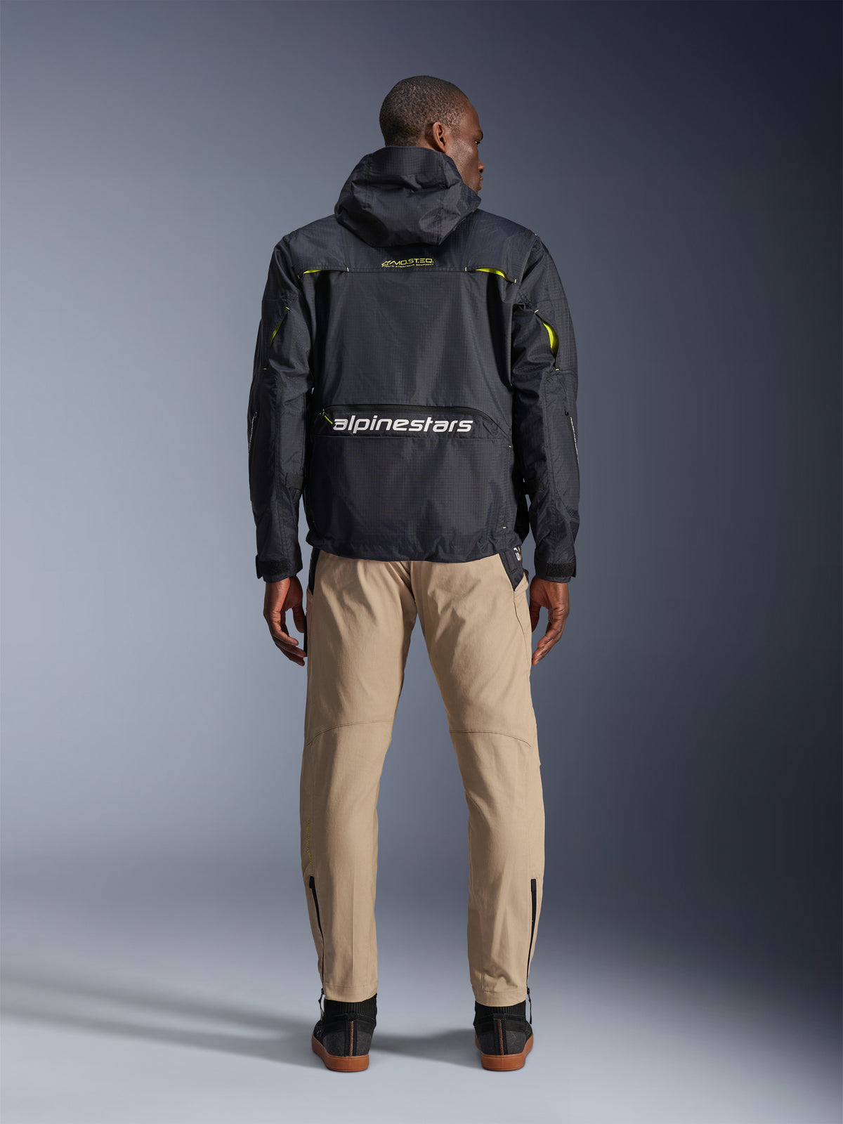 Pantalon Alpinestars Acteon, pantalon de moto urbain, couleur Sable, vu de dos sur un mannequin portant une veste technique noire à capuche et des bottes sombres, doté d'une jambe fuselée avec fermetures éclair aux chevilles et d'une silhouette moderne et épurée.