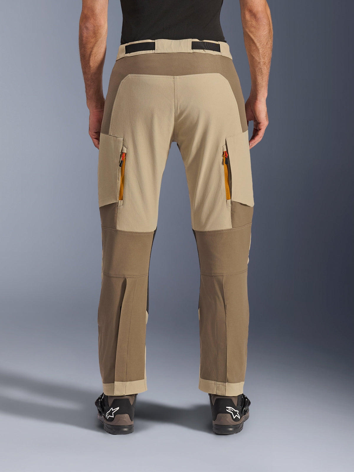 Alpinestars Maxdura Dual Pants, Walnut Aluminum, vue arrière d'un pantalon de moto d'aventure présentant des empiècements beige et tons terre, des évents d'évacuation zippés orange et un design par-dessus la botte, porté avec des bottes Alpinestars noires.