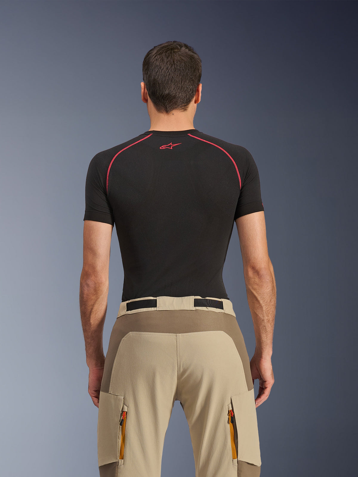 Pantalon Alpinestars Maxdura Dual, pantalon de moto aventure, Walnut Aluminum, vue arrière montrant l'empiècement renforcé à l'assise et le tissu extensible beige, poches cargo zippées avec tirettes orange, porté avec un haut de couche de base Alpinestars noir.