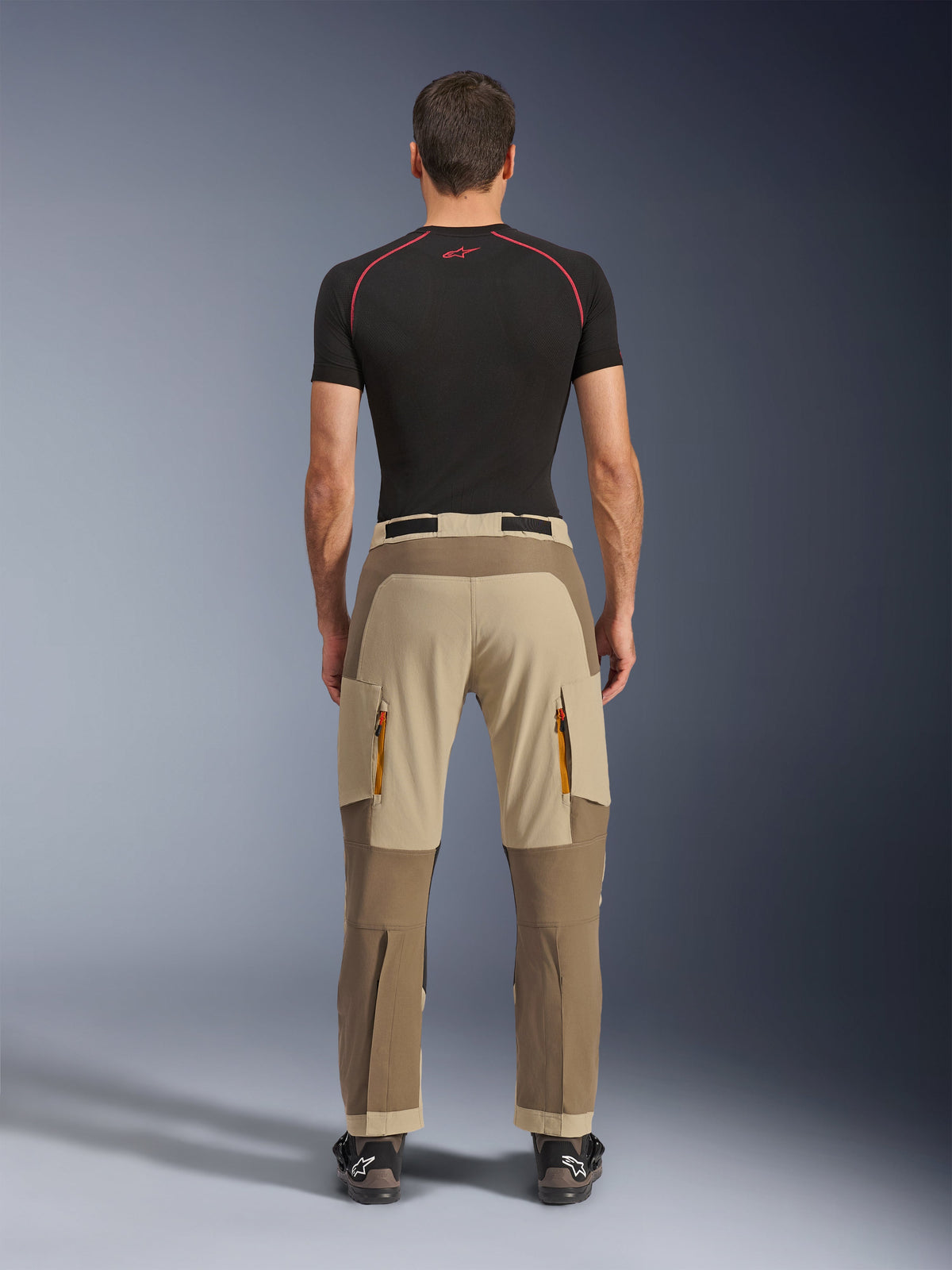 Pantalon Alpinestars Maxdura Dual en Walnut Aluminum, vue arrière montrant un design bicolore marron et beige, pantalon de moto aventure avec poches cargo zippées et assise renforcée, porté avec un haut de compression noir et des bottes de moto.