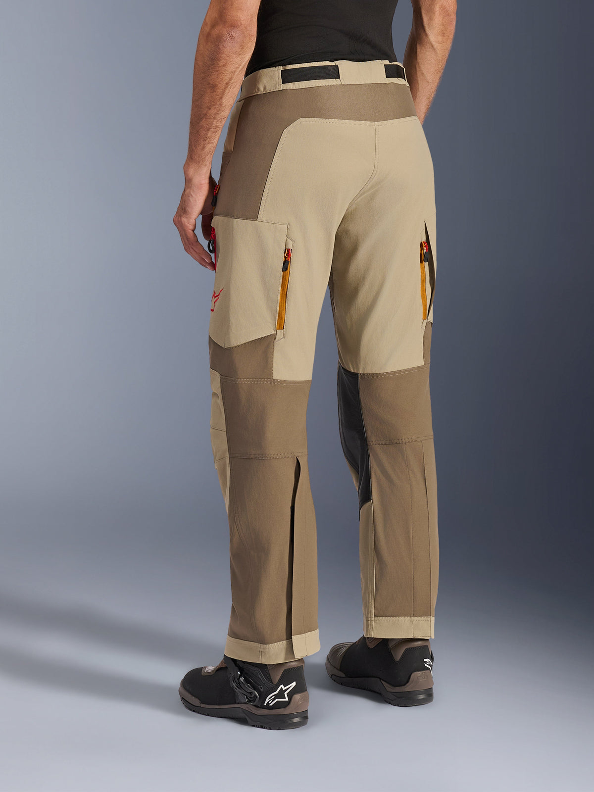 Alpinestars Maxdura Dual Pants en Walnut Aluminum, vue latérale arrière, pantalon de moto d'aventure doté de panneaux beiges et marron, évents d'évacuation arrière zippés, grandes poches cargo et coupe par-dessus la botte porté avec des bottes noires et marron