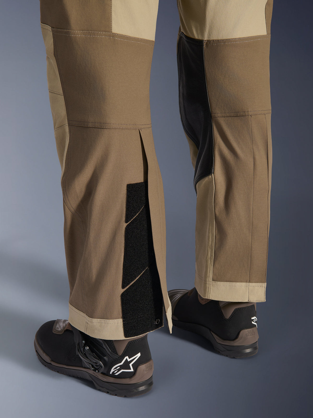 Pantalon Alpinestars Maxdura Dual en Walnut Aluminum, vue détaillée du bas de la jambe et de l'ourlet présentant une conception par-dessus la botte avec fermetures auto-agrippantes réglables et panneaux intérieurs renforcés, associé à des bottes de moto noires.