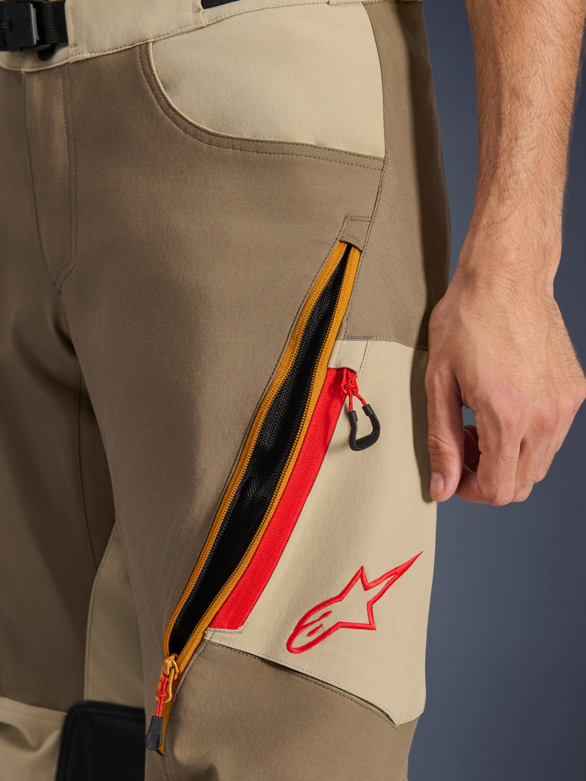 Pantalon Alpinestars Maxdura Dual, Noyer et Aluminium, gros plan sur le détail de la cuisse présentant un large système de ventilation zippé en diagonale avec des accents rouges et un logo Alpinestars rouge sur un panneau beige, équipement de moto d'aventure