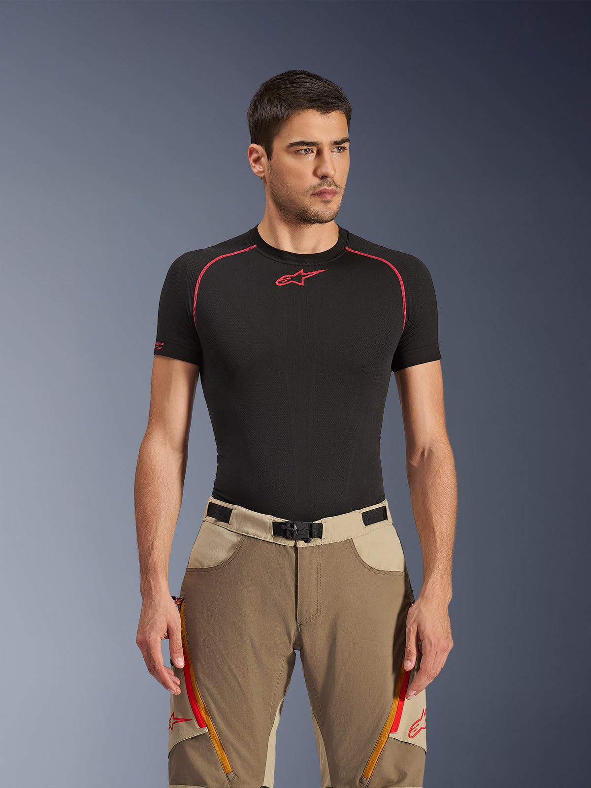 Alpinestars Maxdura Dual Pants en Walnut Aluminum, pantalon de moto aventure beige et marron porté par un mannequin avec un haut technique noir, doté d'aérations zippées orange sur les cuisses, de logos rouges et d'une boucle de ceinture magnétique, portrait en studio.