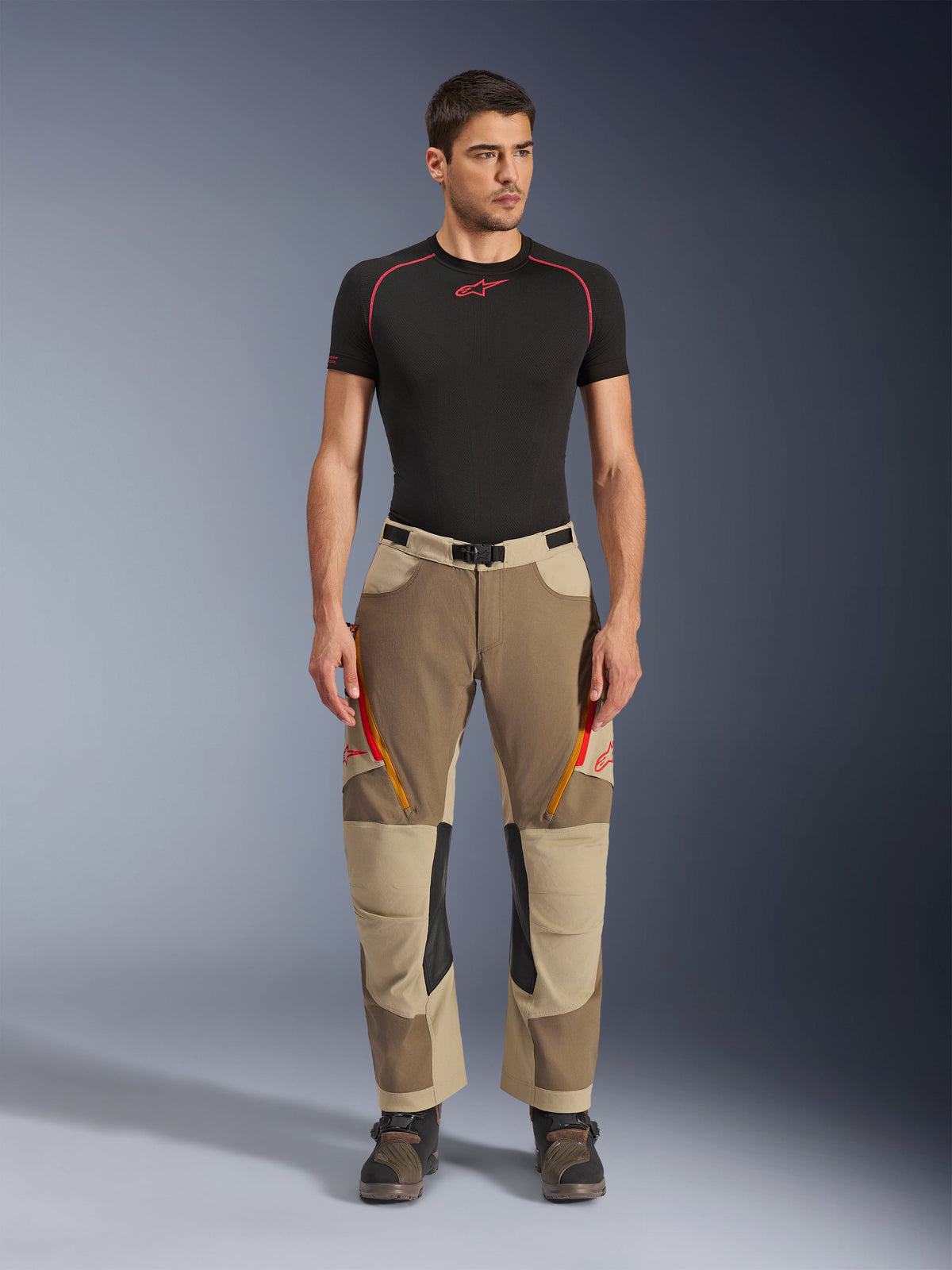 Pantalon Alpinestars Maxdura Dual, pantalon de moto aventure et dual-sport, Walnut Aluminum, porté avec un maillot de compression noir à manches courtes et des bottes d'aventure, doté d'aérations zippées orange et de logos rouges sur les cuisses, ceinture noire avec boucle, design par-dessus la botte pour la pratique de la moto.