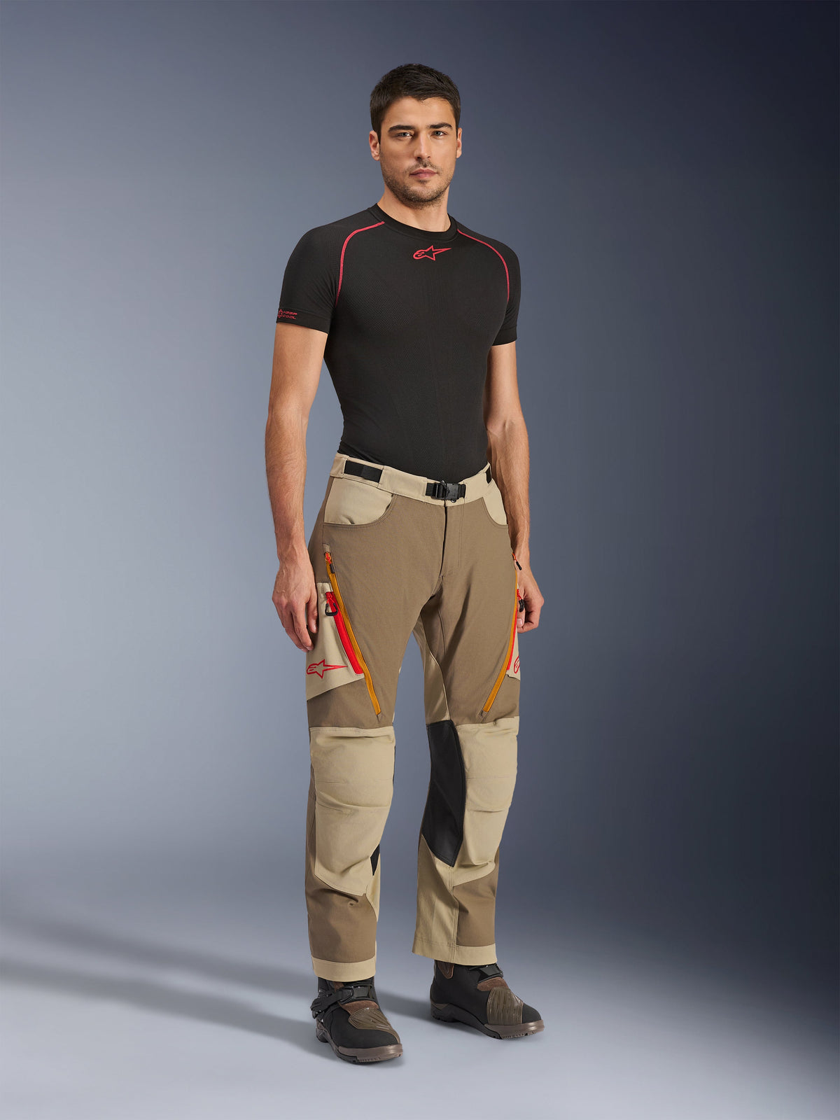 Alpinestars Maxdura Dual Pants en Walnut Aluminum, pantalon de moto d'aventure marron et beige avec fermetures éclair orange et panneaux de genoux en cuir noir, porté avec un t-shirt de base noir et des bottes d'aventure, vue de face en pied