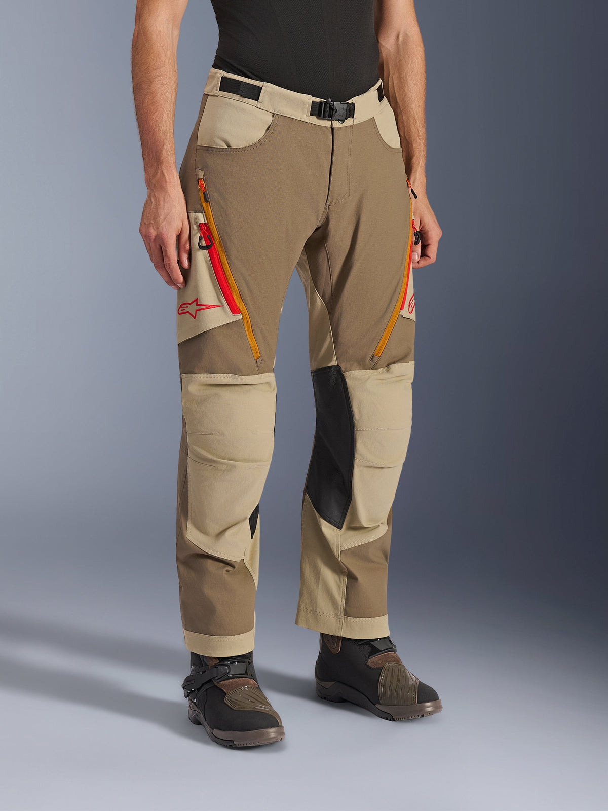Pantalon Alpinestars Maxdura Dual, pantalon de moto d'aventure marron Walnut Aluminum et kaki, doté d'aérations zippées orange et rouge sur les cuisses, de panneaux intérieurs en cuir noir renforcé aux genoux et d'un logo Astars rouge, porté avec des bottes techniques.