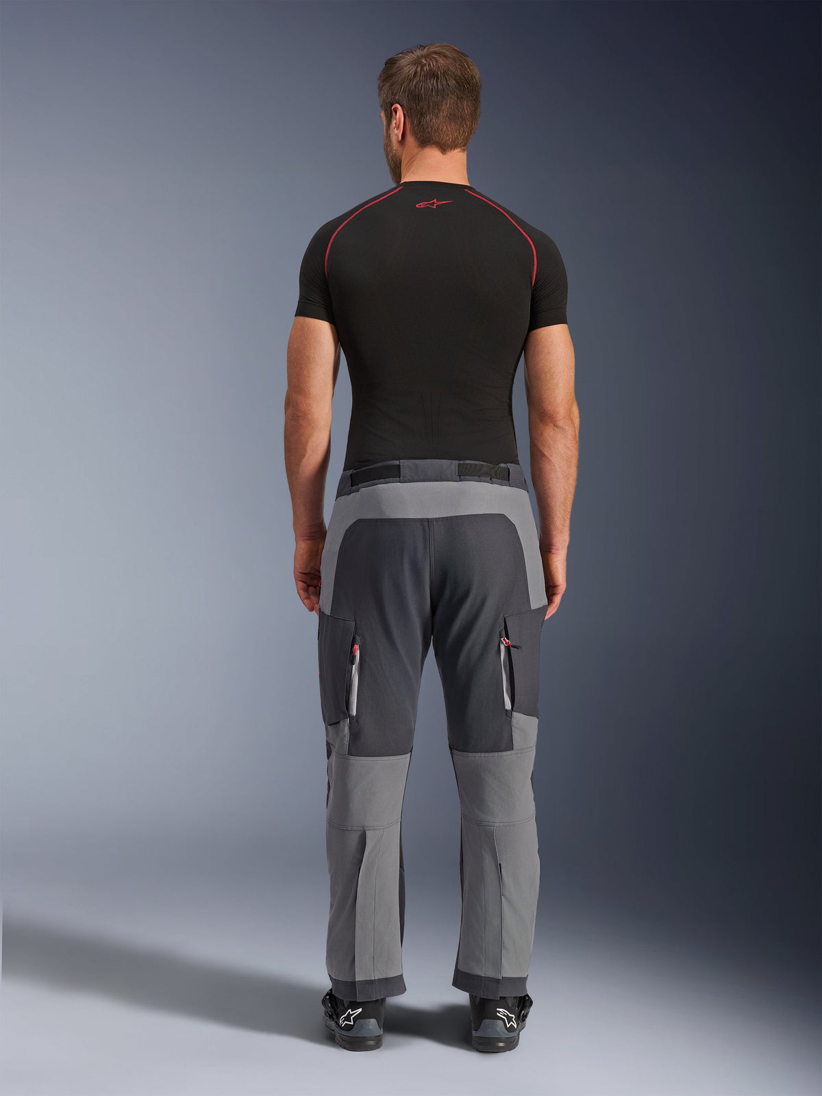 Alpinestars Maxdura Dual Pants, donkergrijze en diepgrijze adventure motorbroek, achteraanzicht op model met zwart T-shirt en laarzen, voorzien van meerkleurige grijze panelen en cargozakken met rits en rode accenten.