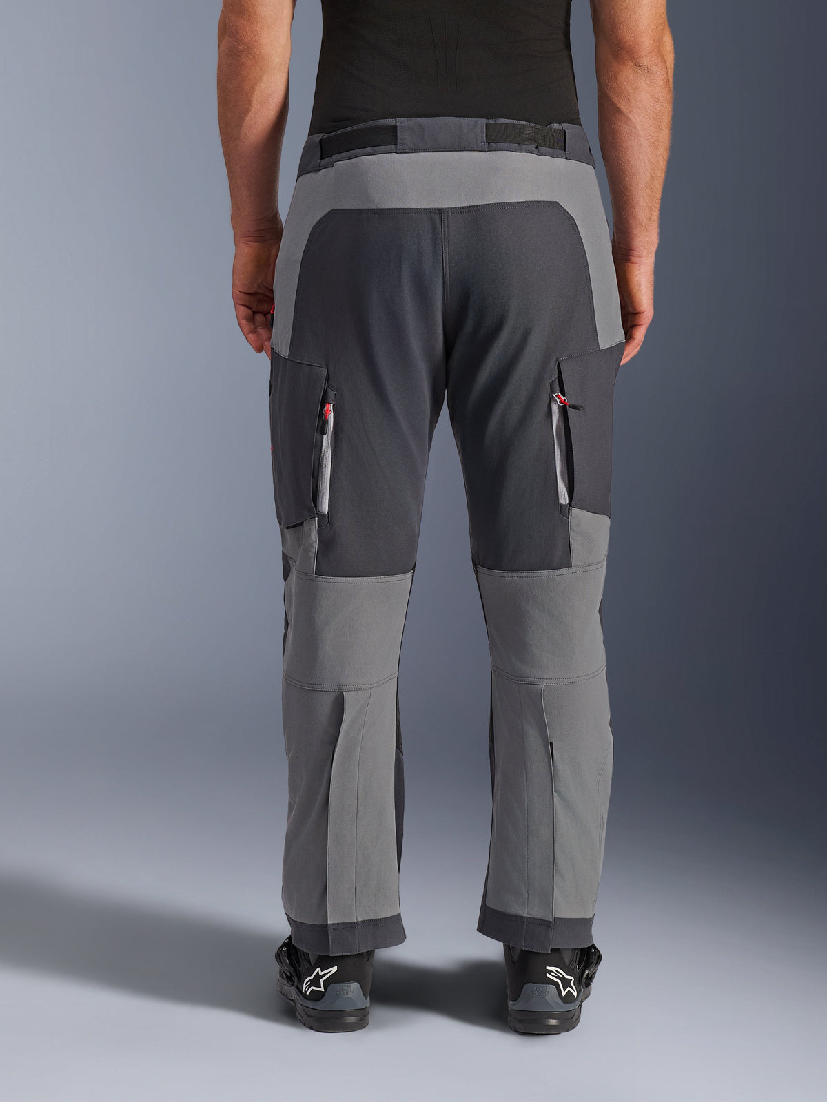 Alpinestars Maxdura Dual Pants, adventure motorbroek, donkergrijs en diepgrijs, achteraanzicht met de nadruk op de verstevigde zitvlak- en kniepanelen, ventilatieopeningen met rits op de bovenbenen, gedragen met zwarte Alpinestars laarzen, robuuste stretch polyamide constructie voor dual-sport rijden.