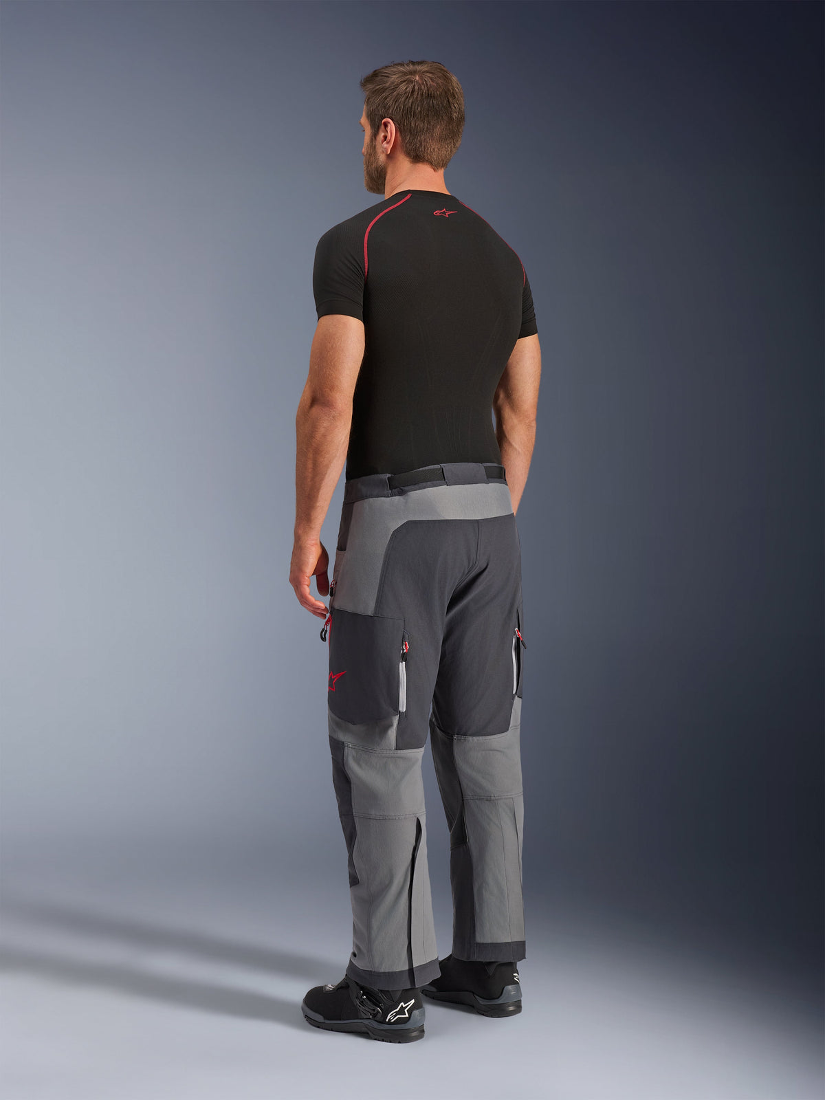 Alpinestars Maxdura Dual Pants, adventure motorbroek, donkergrijs en diepgrijs, gedragen door een model van achteren opzij gezien, met rode accenten op ritsen en logo, versterkte grijze panelen, cargozakken en over-de-laars pasvorm
