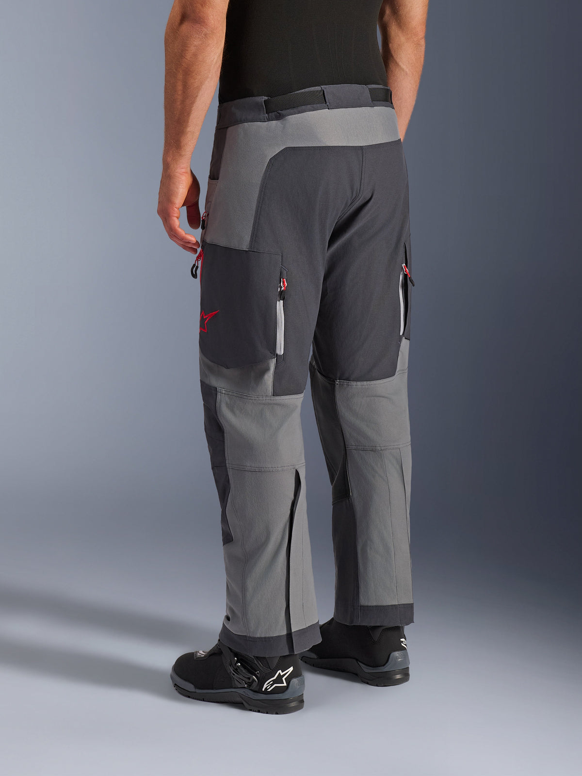 Alpinestars Maxdura Dual Pants, adventure motorbroek, donkergrijs en diepgrijs, achteraanzicht met multi-paneel constructie, rode ritsaccenten en ster-logo, gecombineerd met zwarte Alpinestars laarzen, ontworpen voor dual-sport prestaties