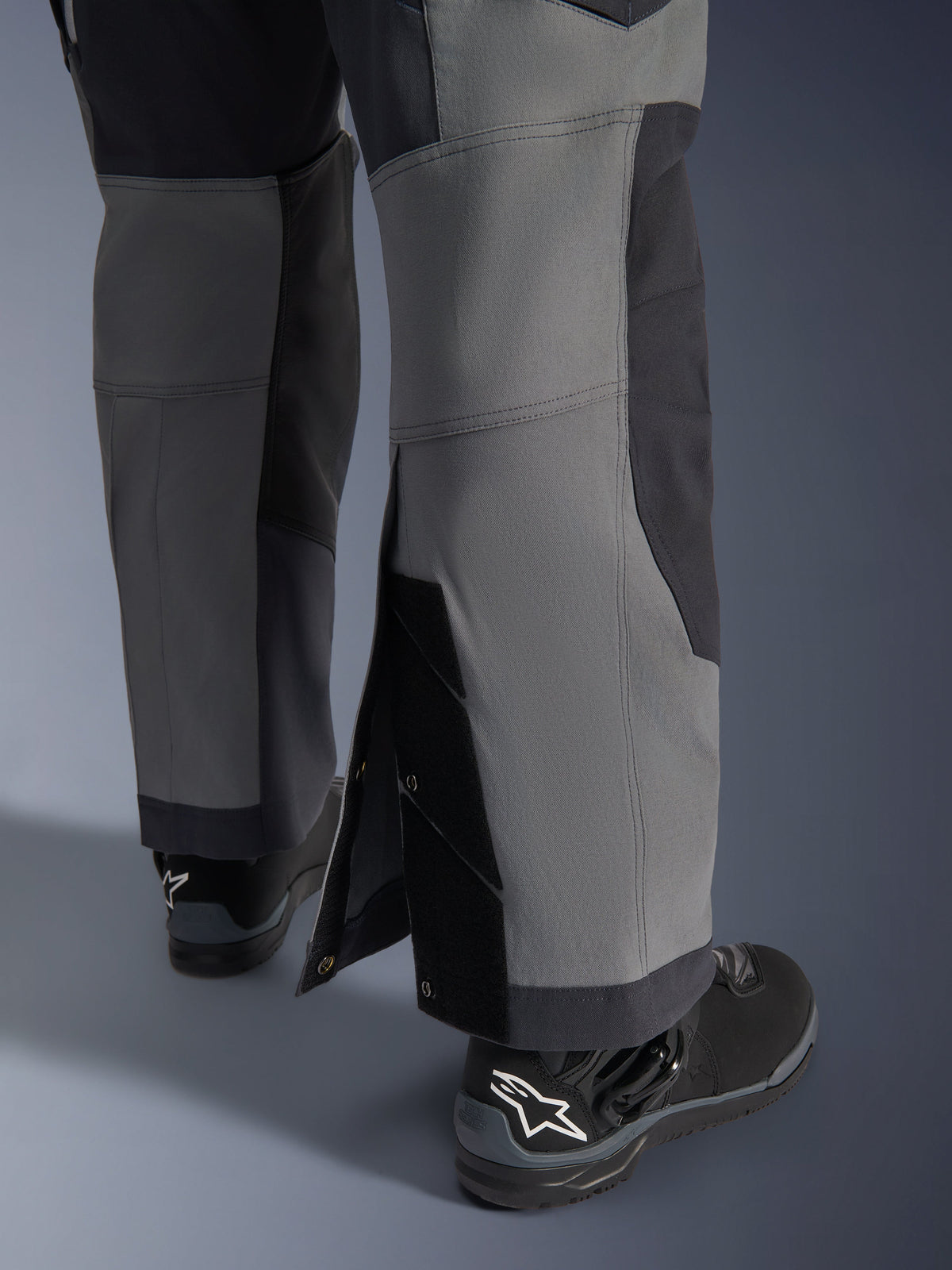 Alpinestars Maxdura Dual Pants, adventure motorbroek, donkergrijs en diepgrijs, close-up van de verstelbare over-de-laars broekzoom met drukknoopsluitingen en verstevigde panelen, gedragen met zwarte Alpinestars laarzen, robuust technisch ontwerp voor dual-sport ritten