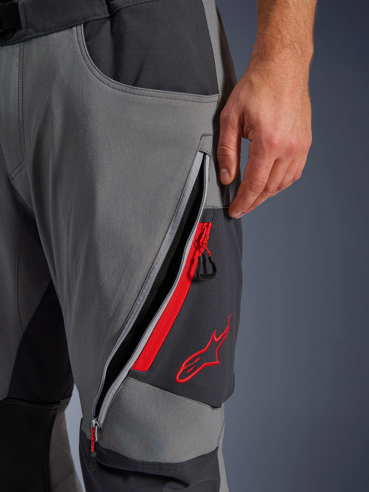 Alpinestars Maxdura Dual Pants, donkergrijze en diepgrijze adventure motorbroek, close-up van open ventilatieopening met rits op het bovenbeen met rode accenten en geborduurd rood sterlogo, technisch ontwerp van stretchstof