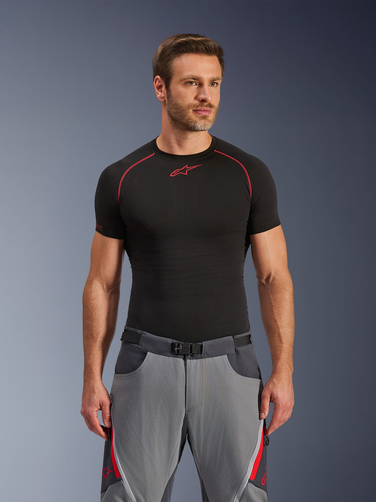 Alpinestars Maxdura Dual Pants, donkergrijze en diepgrijze motorbroek met rode accenten, gedragen met een zwart base layer shirt met rode stiksels, atletische pasvorm met geïntegreerde riem met magnetische gesp, duurzame adventure motorkleding