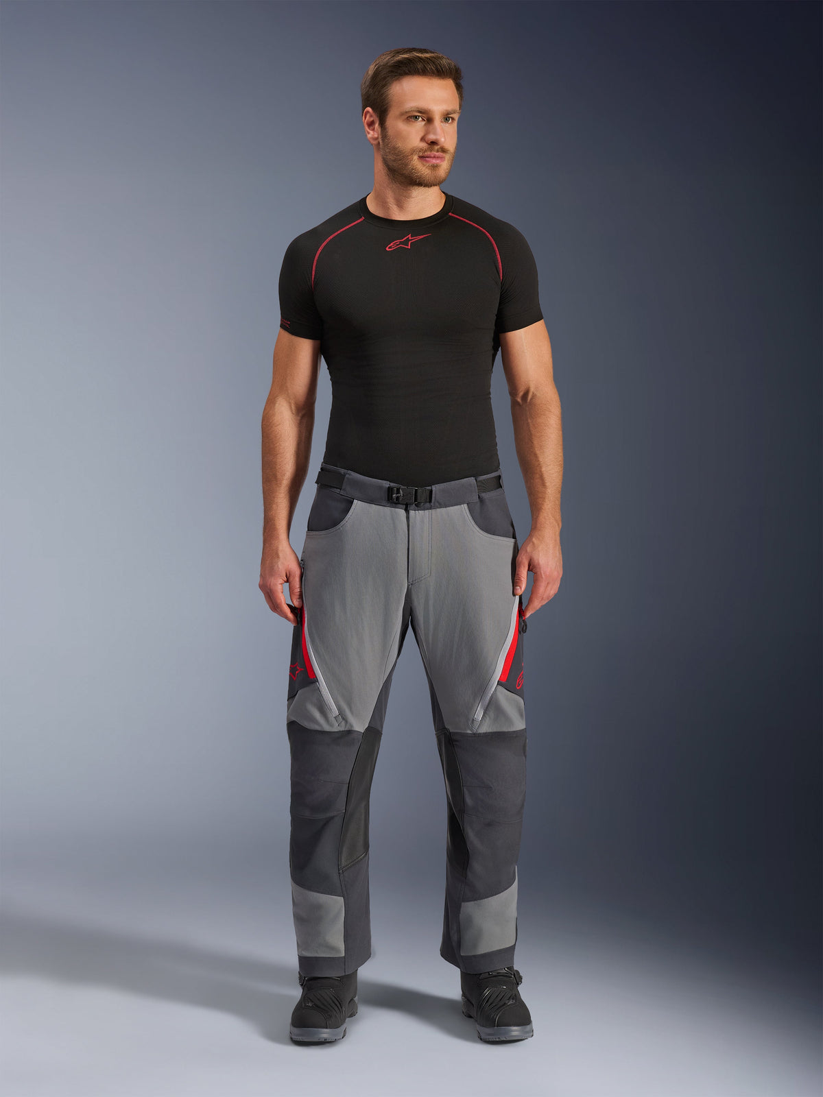 Alpinestars Maxdura Dual broek, motorbroek, donkergrijs en diepgrijs met rode accenten, vooraanzicht gedragen door een model met een zwarte Alpinestars base-layer en zwarte laarzen, voorzien van verstevigde panelen en ventilatieopeningen met rits op de bovenbenen, adventure-stijl motoruitrusting