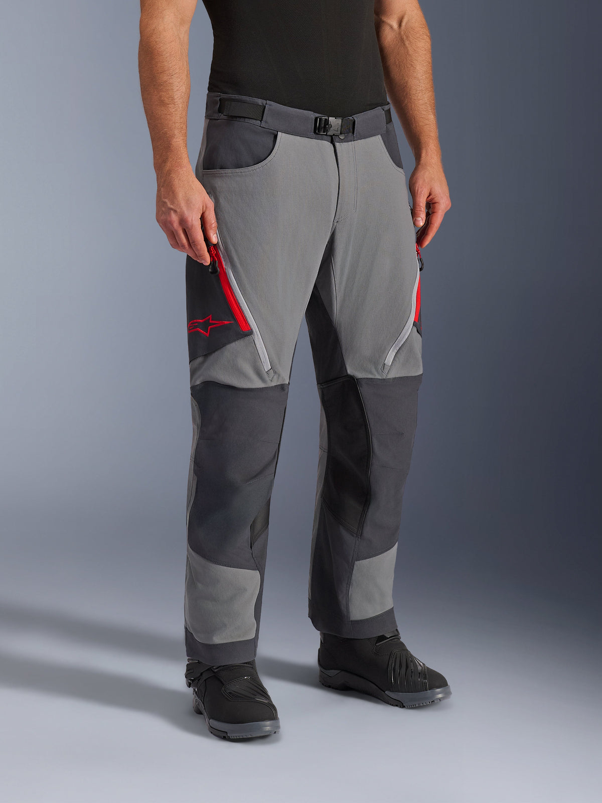 Alpinestars Maxdura Dual Pants, adventure-touring motorbroek in donkergrijs en diepgrijs, met rode ventilatieritsen op de bovenbenen en ster-logo, versterkte donkere panelen op de knieën, gecombineerd met zwarte motorlaarzen