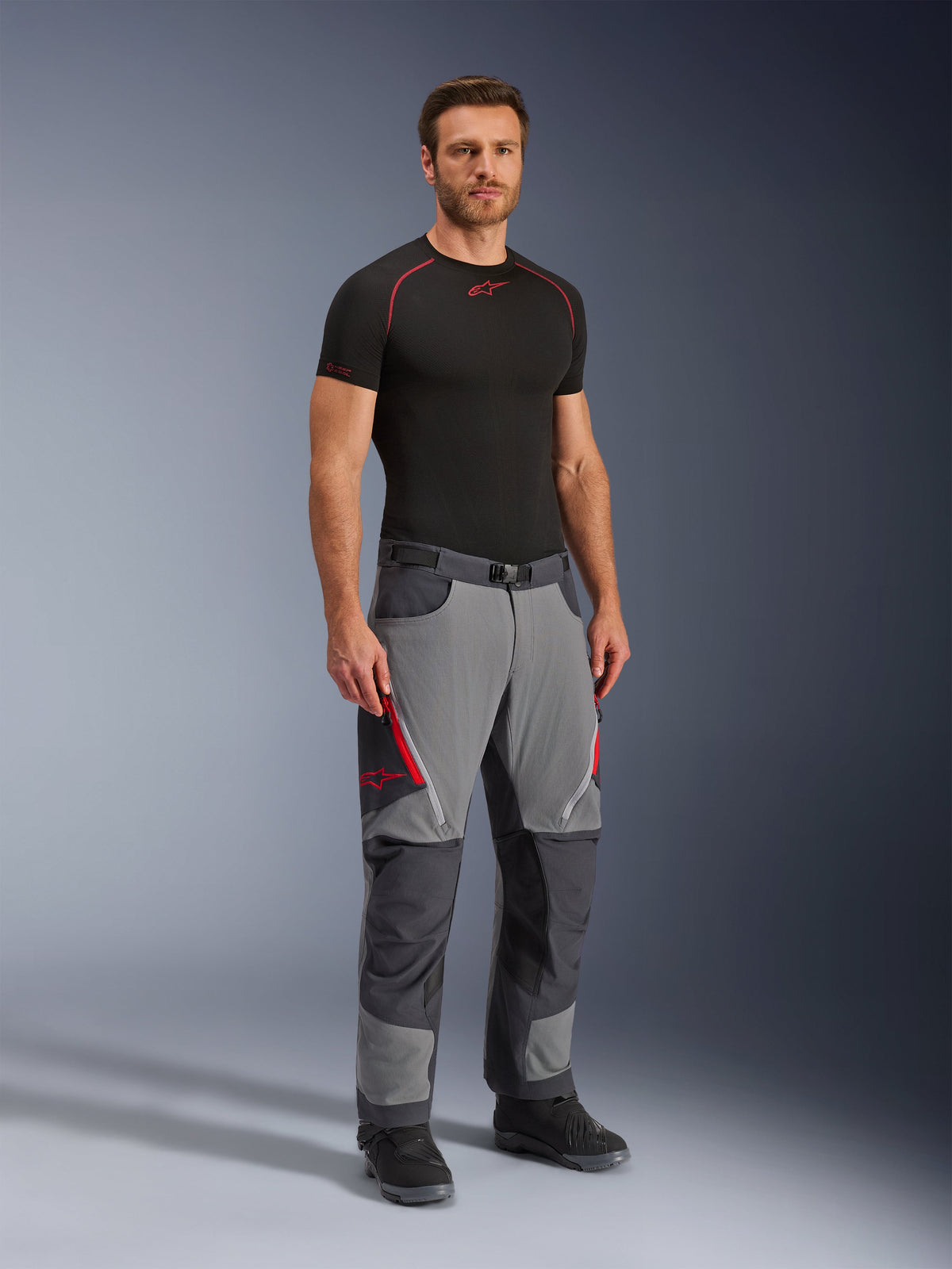 Alpinestars Maxdura Dual Pants, adventure motorbroek, donkergrijs en diepgrijs, gedragen met een zwarte basislaag en laarzen, voorzien van rode ventilatieopeningen met rits, versterkte panelen op de knieën en het zitvlak en een tailleband met riem, sportieve pasvorm voor dual-sport ritten