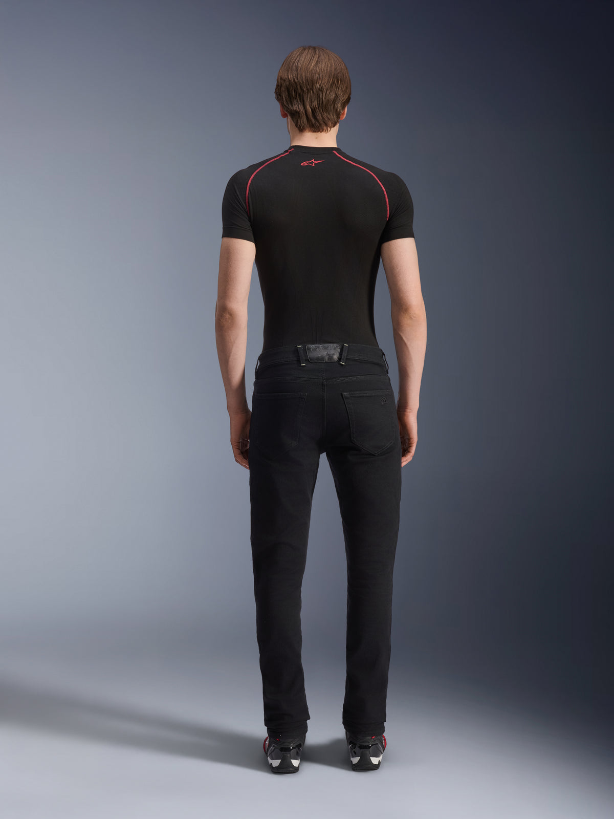 Alpinestars Copper V3 Denim broek in Black Rinse, achteraanzicht op een model, normale motorjeans met rechte pijpen van stretchdenim, gecombineerd met een zwart compressieshirt met korte mouwen en rijschoenen, stedelijke motoruitrusting