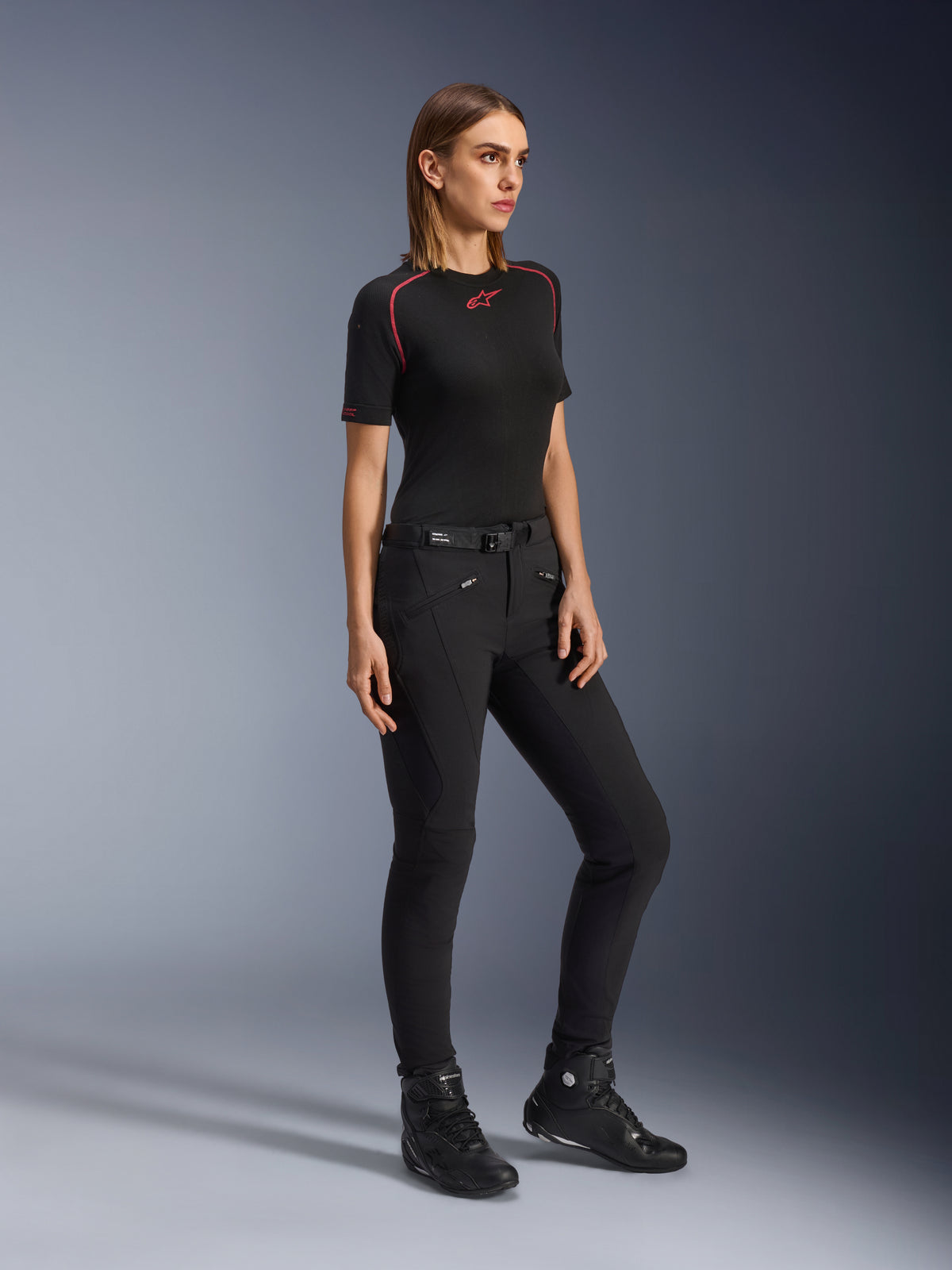 Alpinestars Alamosa Damen-Hose, Slim-Fit-Motorradhose in Schwarz, getragen von einem Modell mit schwarzem Base-Layer und Motorradschuhen, mit Reißverschlusstaschen vorne und einem Taillengürtel mit Magnetverschluss, urbaner Streetwear-Stil.