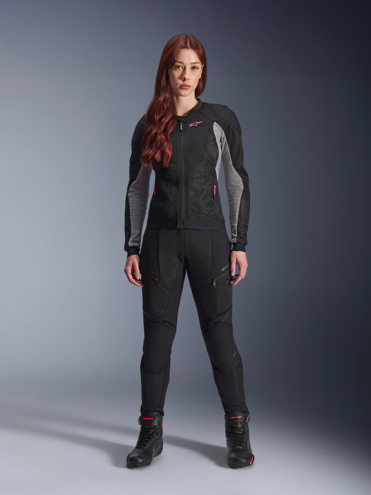 Alpinestars Stella Troop-Air Jacke und Hose, Motorradausrüstung für Damen, Schwarz mit aschegrauen und Pink Fluo Akzenten, getragen von einem weiblichen Model mit roten Haaren, mit Mesh-Belüftung und schützendem Design, stehende Pose im Studio