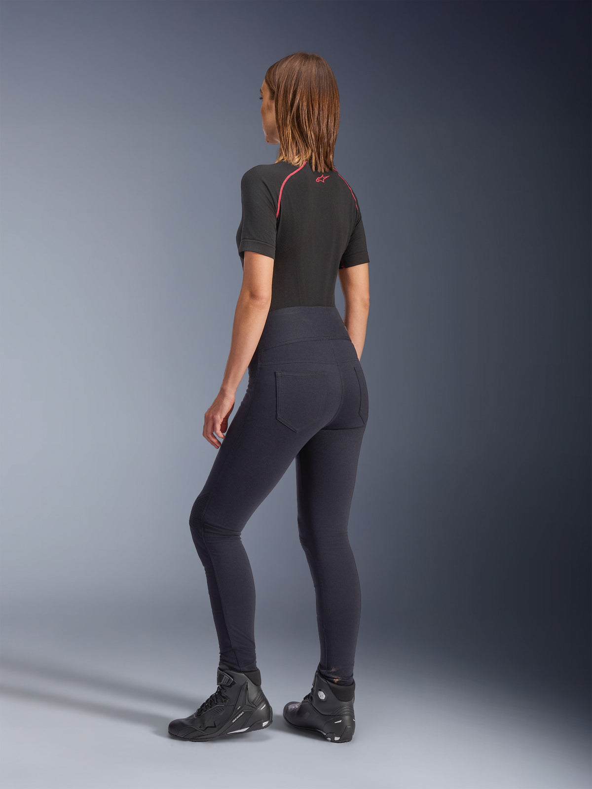 Alpinestars Stella Flex-Ast dameslegging, zwart, achteraanzicht met hoge taille en achterzakken, gedragen met een zwarte basislaag met korte mouwen en rode accenten en zwarte motorschoenen, urban eigentijdse motorkleding