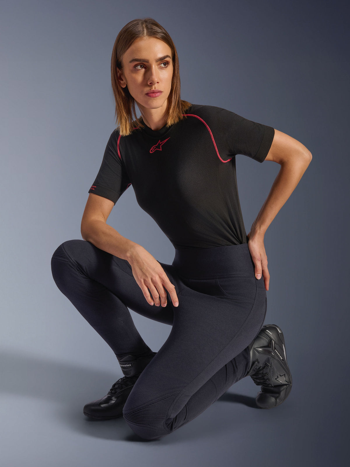 Alpinestars Stella Flex-Ast Leggings, zwart, gedragen door een vrouw knielend in een studio, gecombineerd met een zwarte performance-top met korte mouwen en roze accenten en zwarte motorlaarzen, voorzien van een hoge taille en flexibele panelen voor stedelijke ritten.