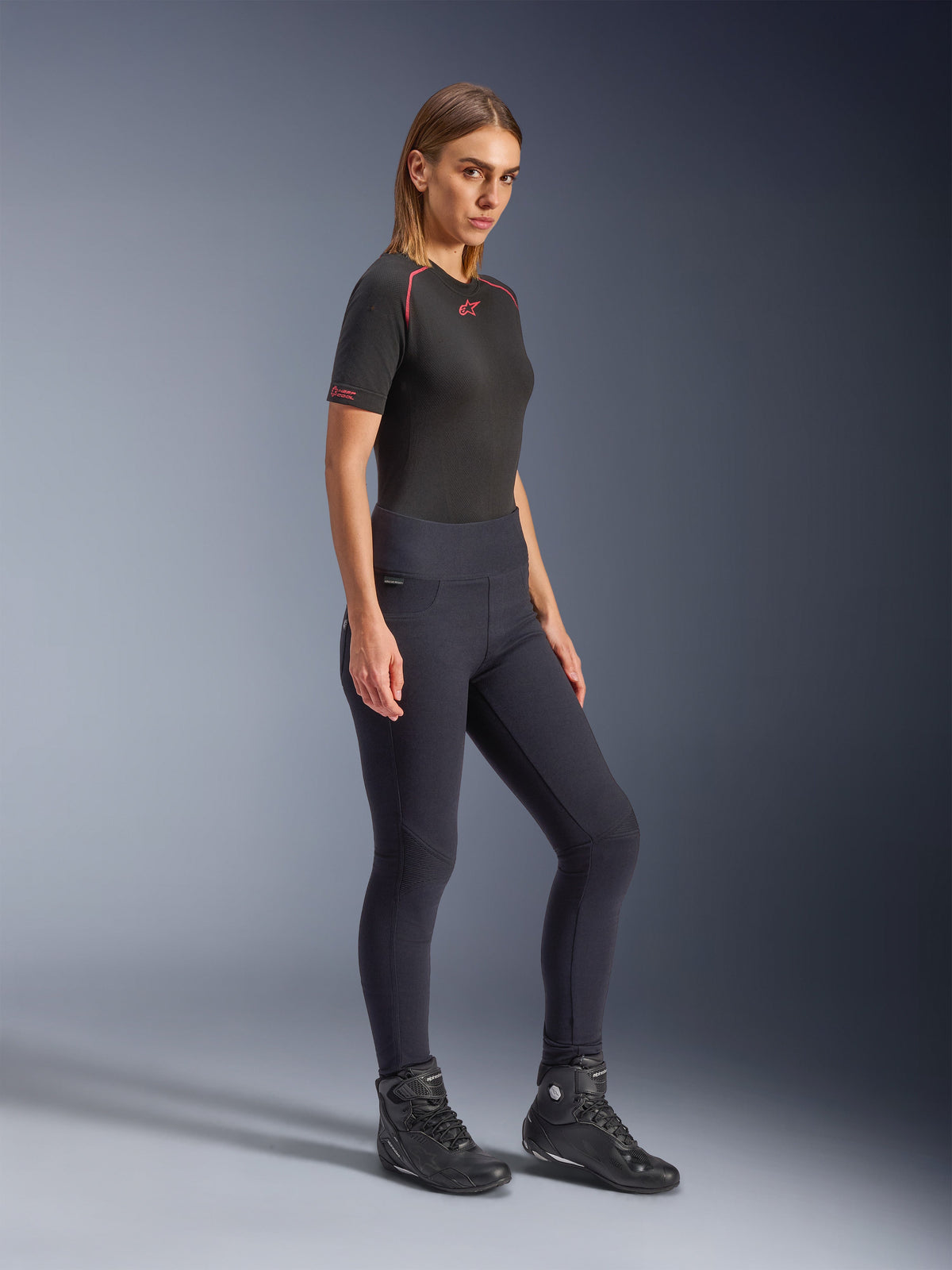 Alpinestars Stella Flex-Ast Leggings, motorleggings voor dames in het zwart, high-waisted slim-fit ontwerp, gedragen met een zwart T-shirt met rode accenten en zwarte motorlaarzen, volledige lichaamsopname in studio.