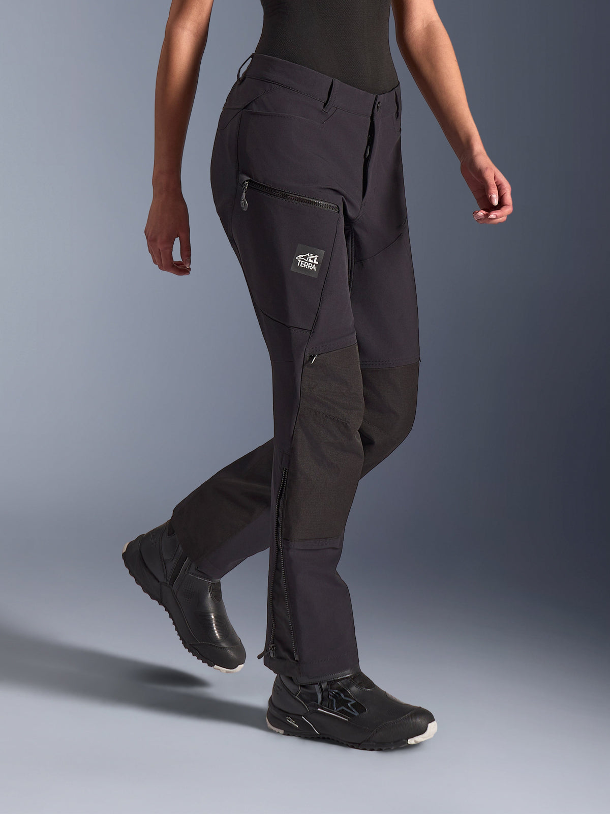 Pantalon Stella Flex-AST Explorer Alpinestars pour femme, noir, pantalon de moto d'aventure, doté d'une construction en textile extensible avec renforts ripstop aux genoux, poches cargo zippées avec marquage TERRA et fermetures éclair au bas des jambes, porté avec des chaussures de moto noires.