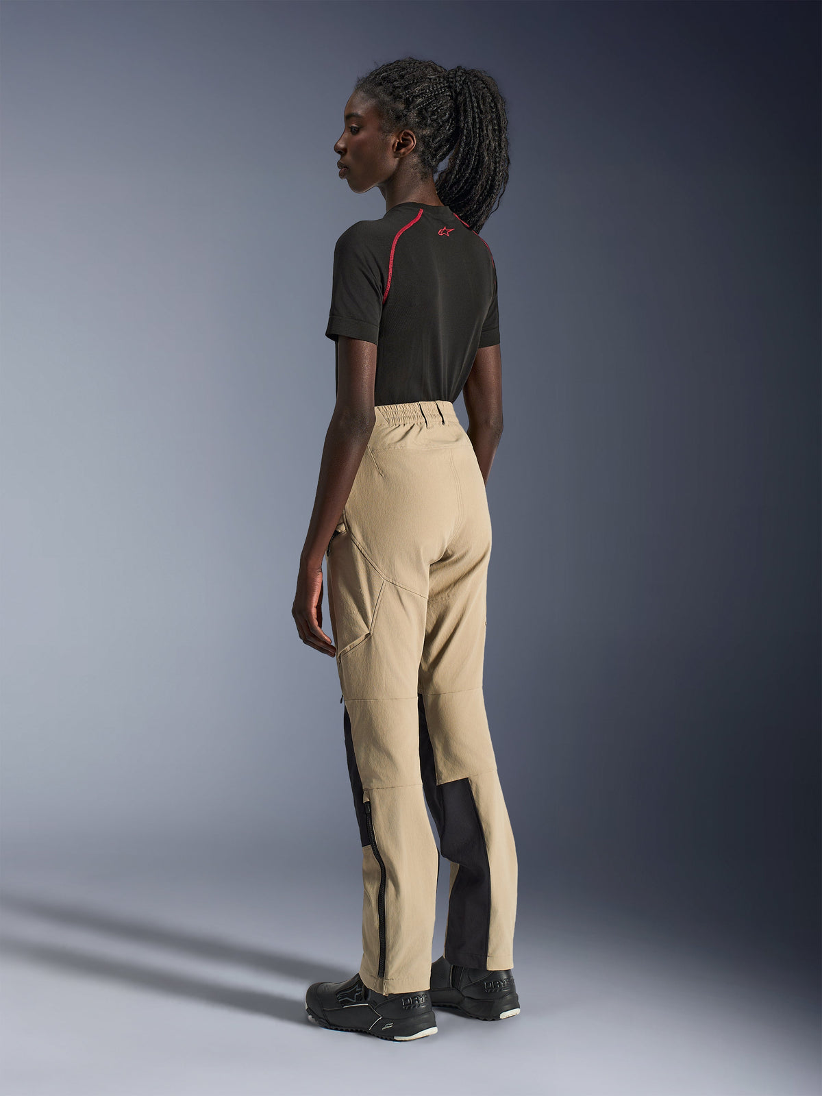 Alpinestars Stella Flex-AST Explorer broek, dames motorbroek, zandkleur en zwart, zij-achteraanzicht op een model in combinatie met een zwart t-shirt en motorlaarzen, toont ripstop-verstevigingen en ritsopeningen aan de onderbenen.