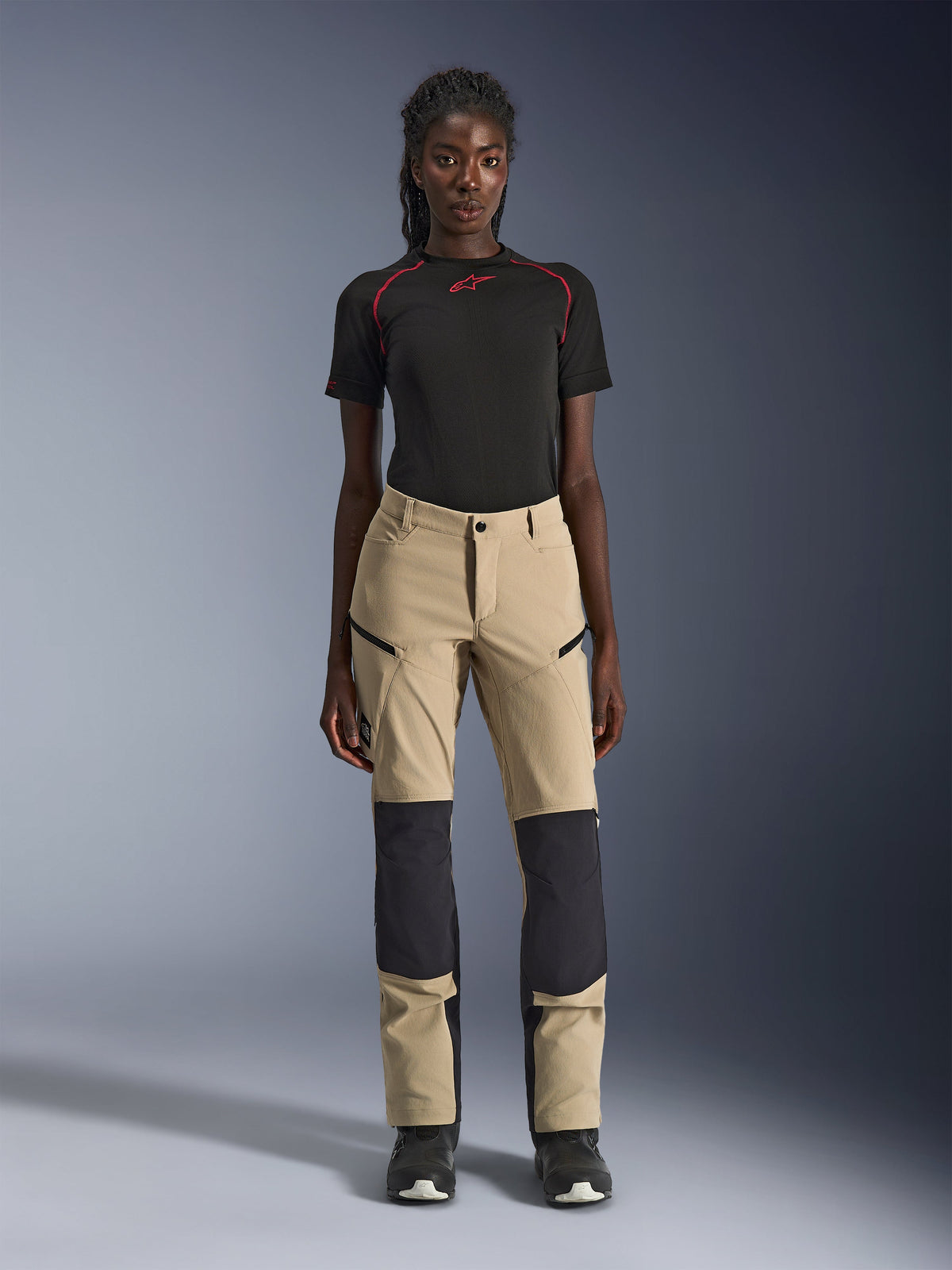 Alpinestars Stella Flex-AST Explorer damesmotorbroek, beige met zwarte panelen, gedragen door een model met een zwart shirt met korte mouwen en zwarte laarzen, voorzien van verstevigde kniepanelen, cargozakken met ritssluiting en ademende ripstop-stof voor adventure-ritten in warm weer.