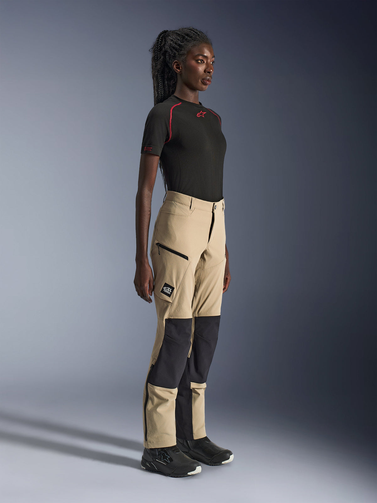 Alpinestars Woman Stella Flex-AST Explorer Pants, zandkleurige en zwarte adventure touring broek, zijprofiel van een model dat een zwart Alpinestars t-shirt en motorlaarzen draagt, met ripstop knieverstevigingen en cargozakken met ritssluiting.