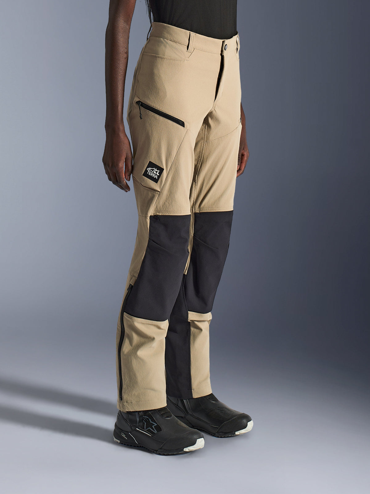 Vrouw draagt Alpinestars Stella Flex-AST Explorer Pants, zandkleurige en zwarte adventure motorbroek met zwarte ripstop kniepanelen, cargozakken met ritssluiting, 'ALL TERRA' logo patch en ritsen aan de onderkant van de pijpen.