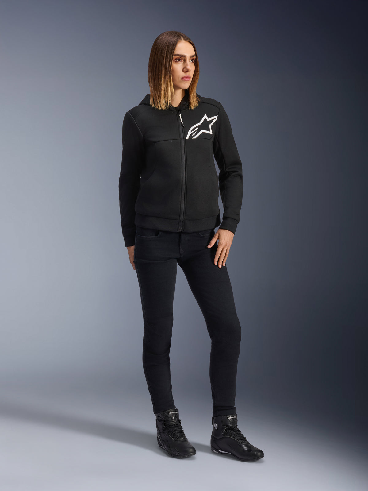 Femme portant le jean de moto Daisy V3 Femme Alpinestars, pantalon de moto coupe slim en Rinse Black, associé à un sweat à capuche zippé noir et des bottes de moto, équipement de protection urbain décontracté