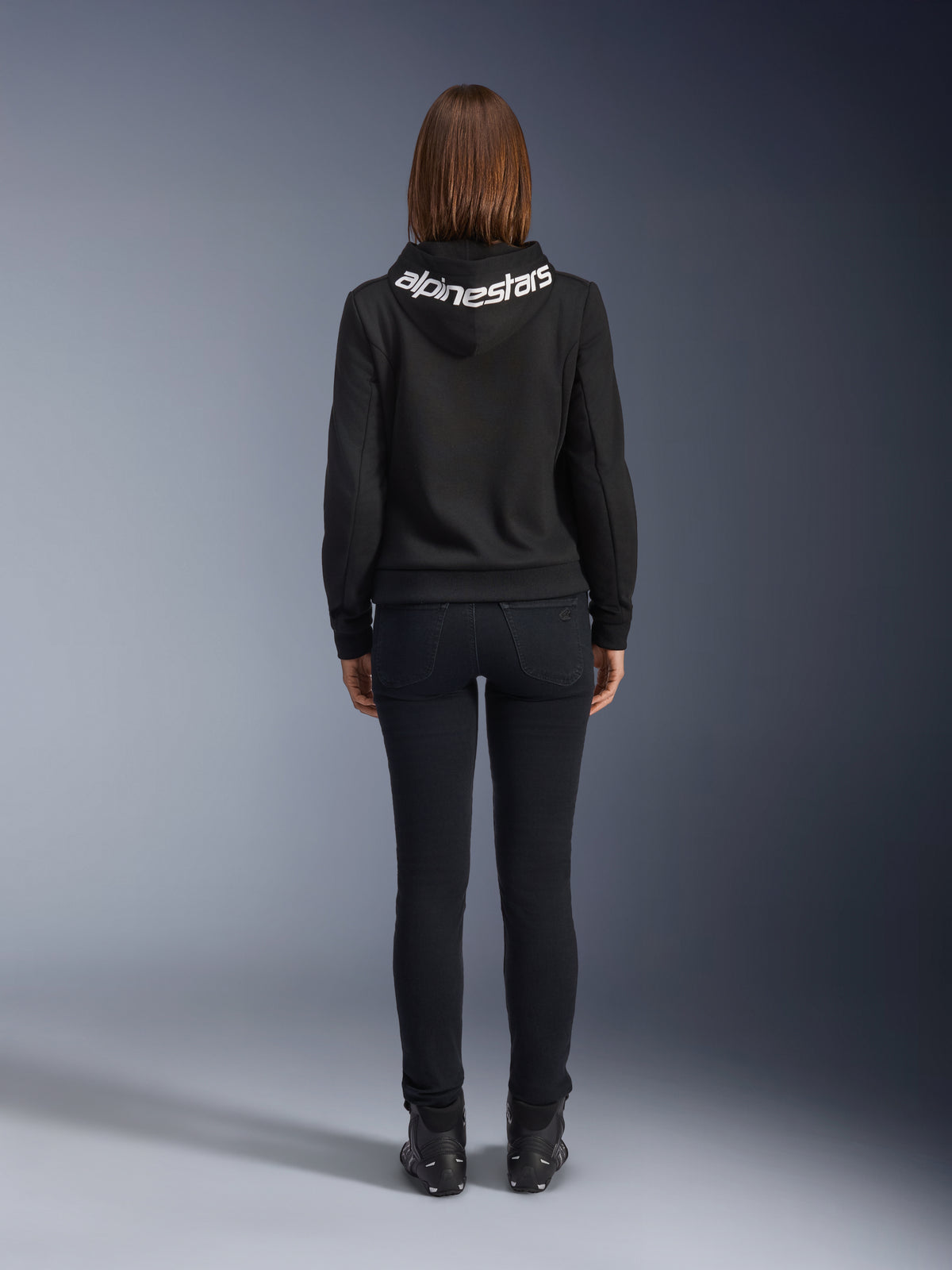 Denim de moto pour femme Daisy V3, Black Rinse, pantalon de moto coupe slim, vue arrière montrant la taille haute et une silhouette féminine, associé à un sweat à capuche Alpinestars noir et des bottes de moto noires, équipement de conduite urbain décontracté