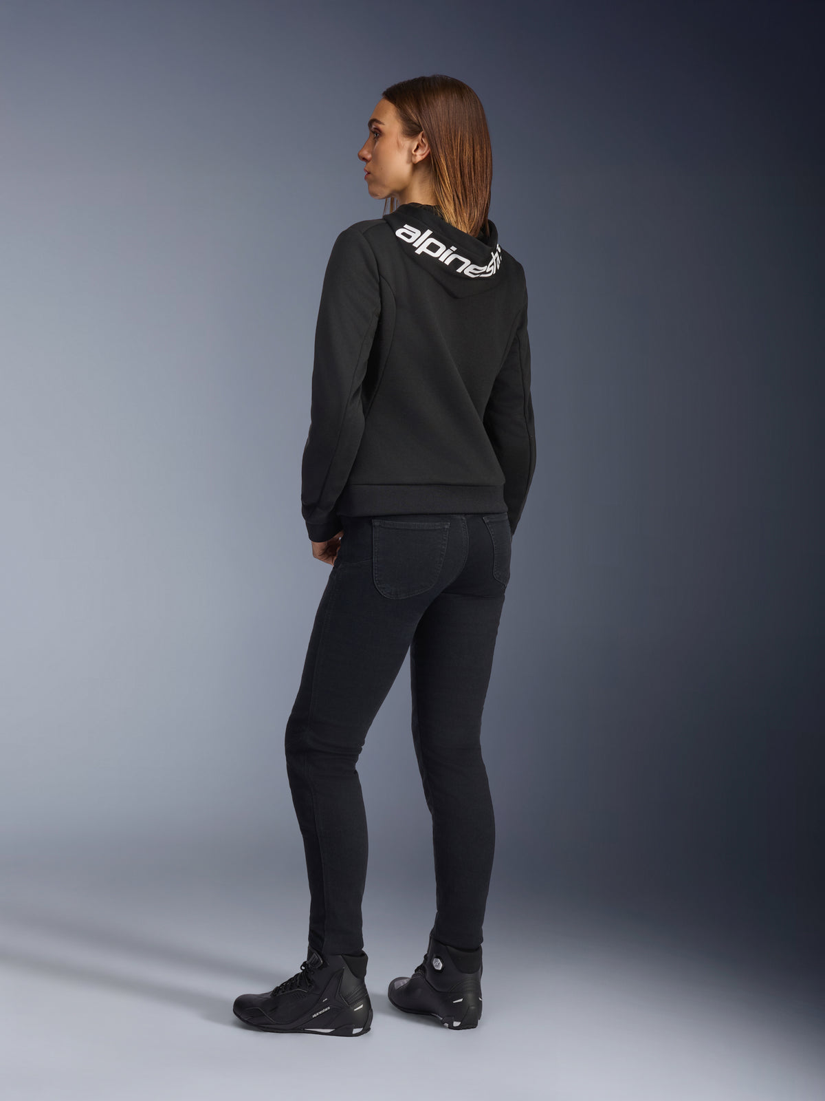 Alpinestars Daisy V3 Women's Riding Denim, pantalon de moto pour femme, noir, vue arrière montrant la coupe ajustée et la taille haute, associé à un sweat à capuche Alpinestars noir et des bottes de pilotage pour un look moto urbain