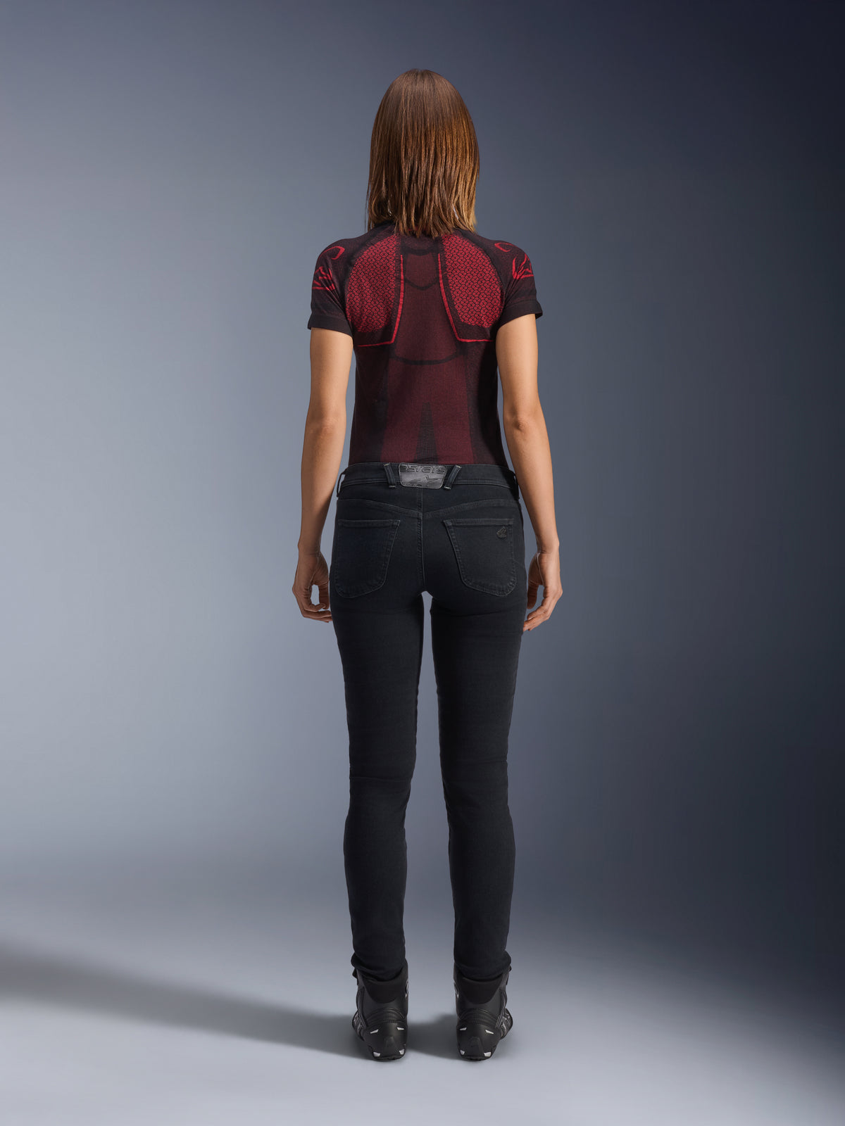 Alpinestars Daisy V3 Women's Riding Denim, Noir Rinse, pantalon de moto coupe slim vu de dos, doté d'une taille haute et de poches arrière, associé à un haut technique à motifs rouge et noir et des bottes noires, équipement de moto urbain pour femme