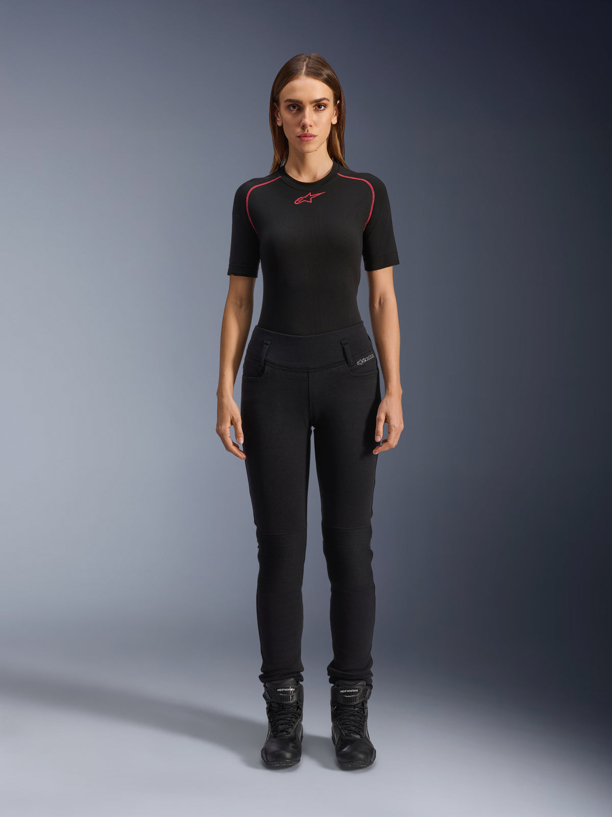 Alpinestars Banshee Damen-Leggings, schwarze Motorrad-Leggings, figurbetonte anatomische Passform, getragen von einem weiblichen Model mit schwarzem Baselayer und Motorradstiefeln, mit gerippten Knieeinsätzen und hoch geschnittenem Bund für urbane Fahrten.