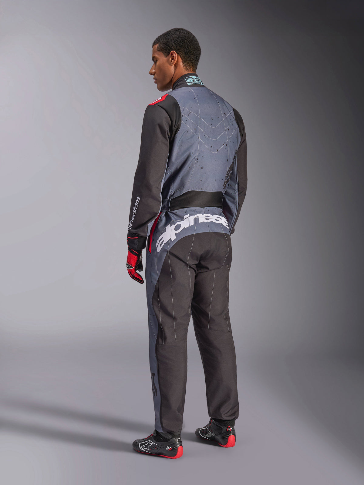 Vue arrière d'un mannequin portant la combinaison Alpinestars KMX-9 V3 Graphic 6, combinaison de karting, Anthracite Noir Rouge, avec un logo Alpinestars blanc distinctif au bas du dos, un design à panneaux noirs et gris avec des accents rouges, portée avec des gants et des chaussures de course assortis.