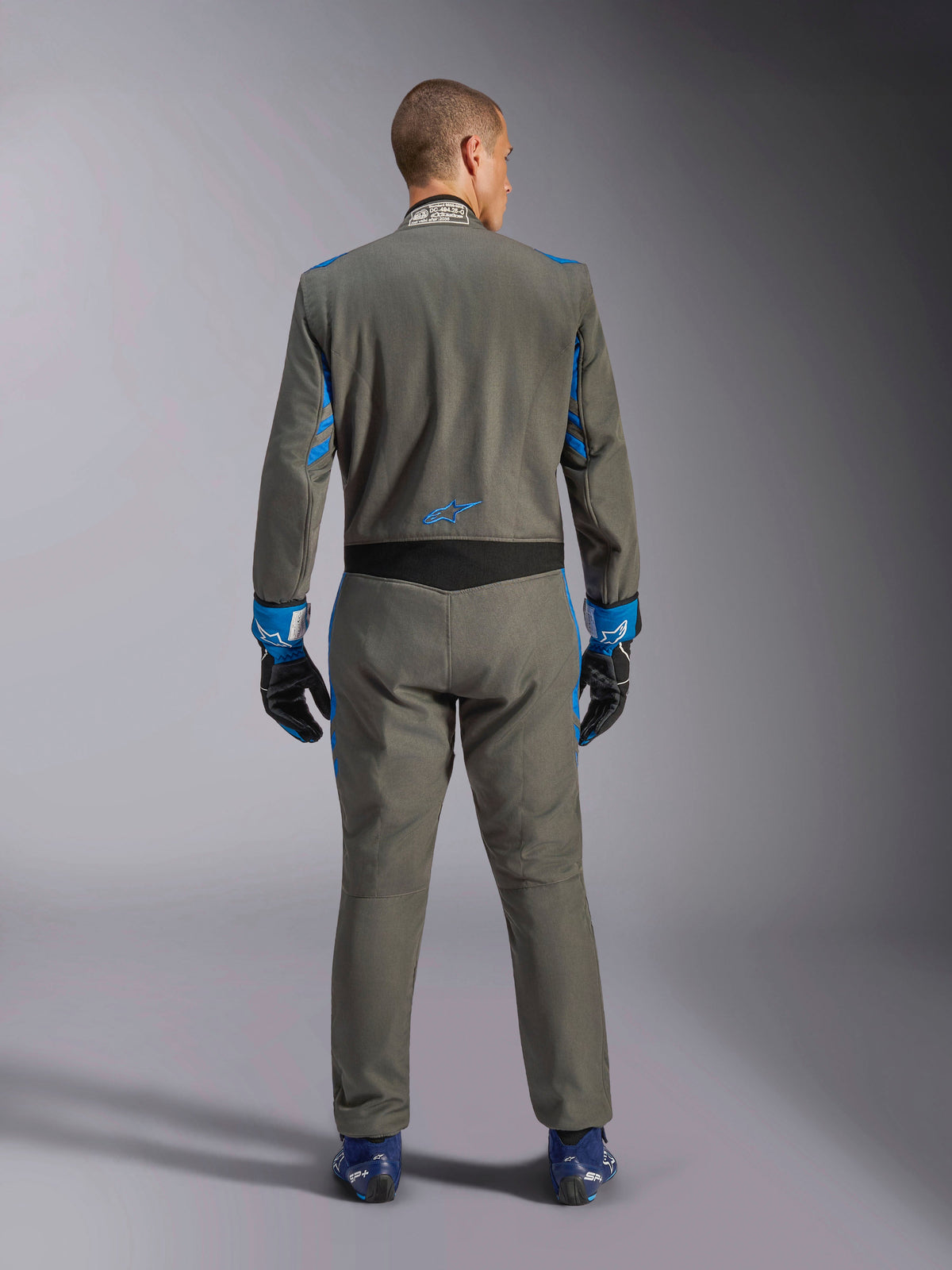 Alpinestars GP Race V3 Anzug FIA, Rückansicht, Anthrazit-Blau mit leuchtend blauen Details an Armen und Schultern, mit blauem Stern-Logo auf dem unteren Rücken, getragen mit passenden Rennhandschuhen und blauen Schuhen, professioneller Auto-Rennanzug