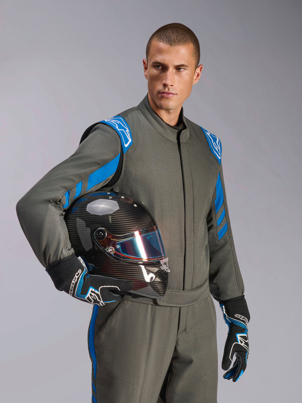 Männlicher Rennfahrer im Alpinestars GP Race V3 Anzug FIA in Anthrazit-Blau, hält einen Carbon-Rennhelm mit schillerndem Visier, professionelle Autorenn-Ausrüstung mit blauen Schulterakzenten und passenden Rennhandschuhen.
